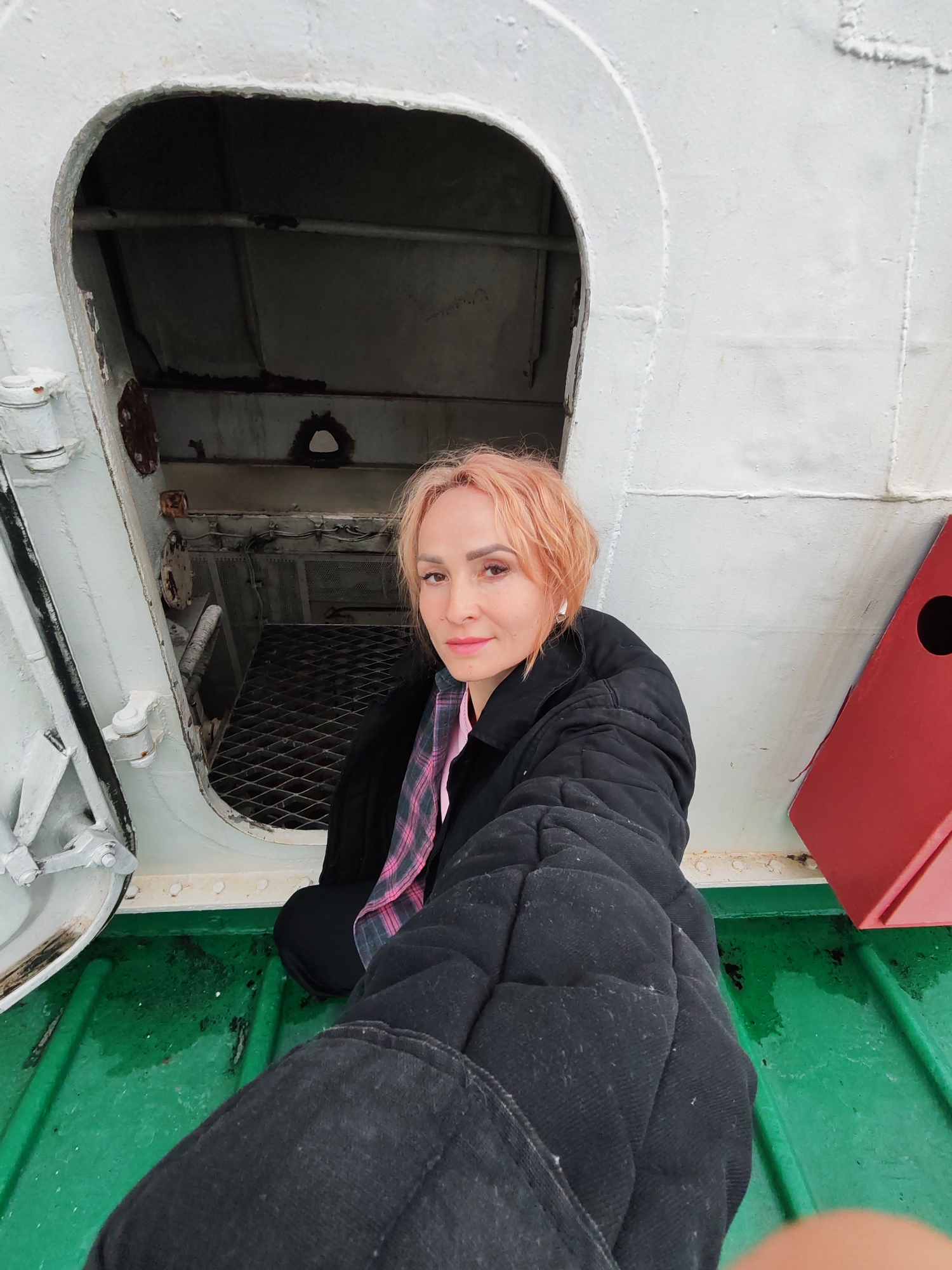 A woman with short, peachy-pink hair and a pale complexion is taking a selfie on a green, metallic surface, possibly a ship's deck, in Ozernoe Ust'e, Russia. She is wearing a black, quilted jacket over a pink and grey plaid shirt. Her left hand is extended towards the camera, obscuring part of the foreground. Behind her, an open, arched doorway reveals a dark, gridded interior. The exterior of the doorway and the surrounding structure are painted white. To the right of the woman, a red, box-like object is visible against the white wall. The green surface appears wet and somewhat weathered.