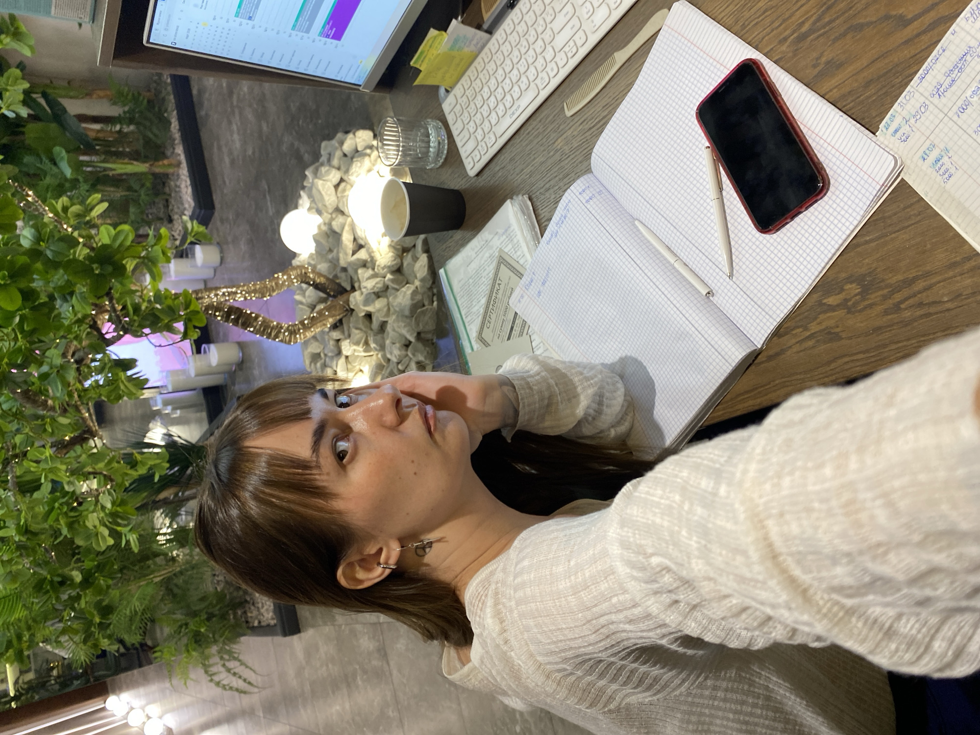 The image shows a woman in a white sweater sitting at a desk. The desk is covered with various items including a computer monitor, keyboard, notebooks, a black phone, pens, a coffee cup, and a glass. The woman is looking at the camera. In the background, there are plants and decorative lights. The certificate visible in the image indicates it's worth 5000 rubles. OCR text "27.03 31 03-3000/02<8 ych 1 29.03 0229 10йсии" might point to Votkinsk, Russia.