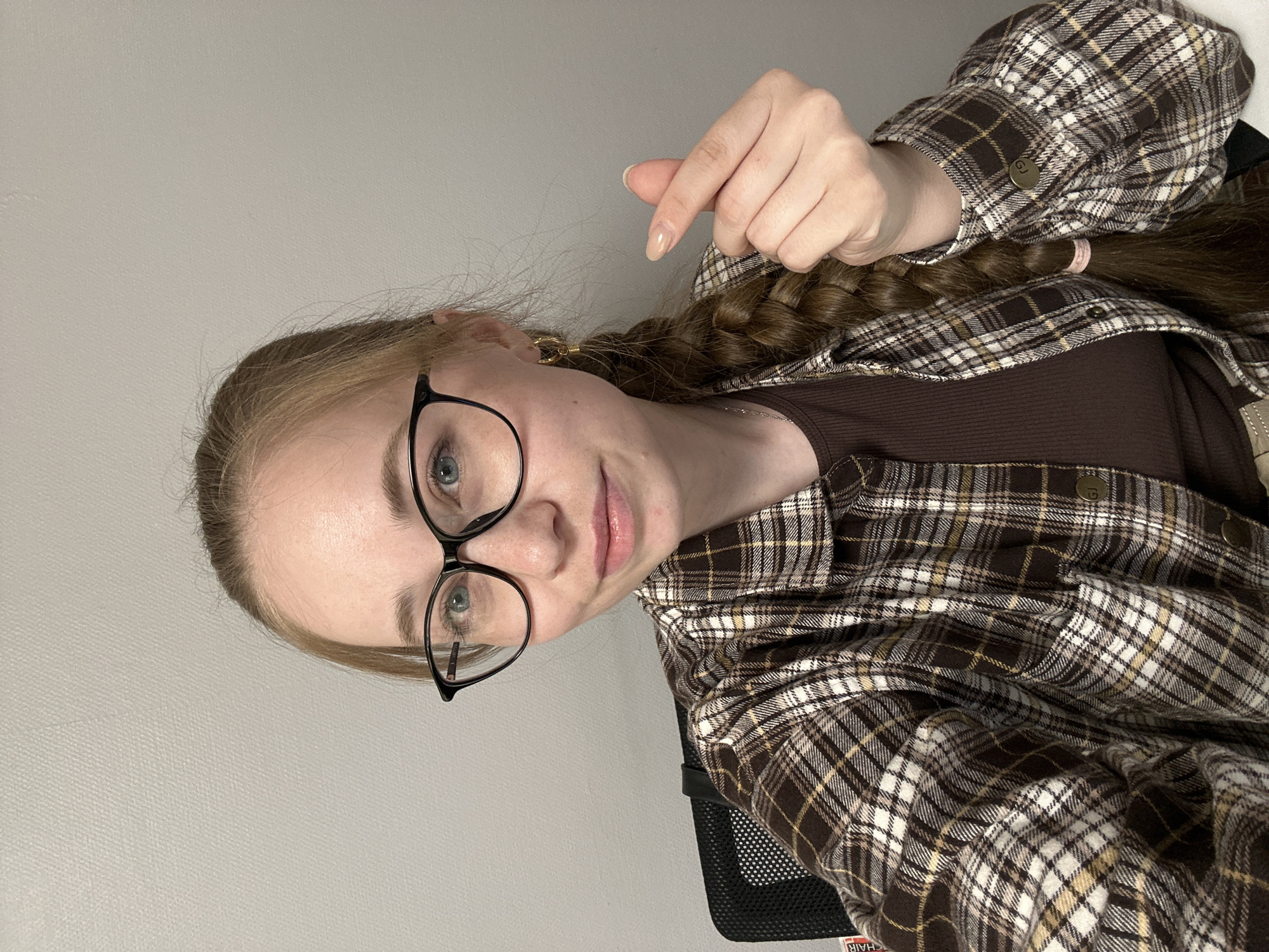 A fair-skinned woman with long blonde hair braided behind her is visible from the chest up. She is wearing black-framed glasses, a brown ribbed shirt, and a brown and white plaid flannel shirt. She is making a finger heart gesture with her right hand. The background is a plain gray wall. The woman is seated in a black mesh chair. No information is available to determine if the location is the city of Saint Petersburg, Russia.