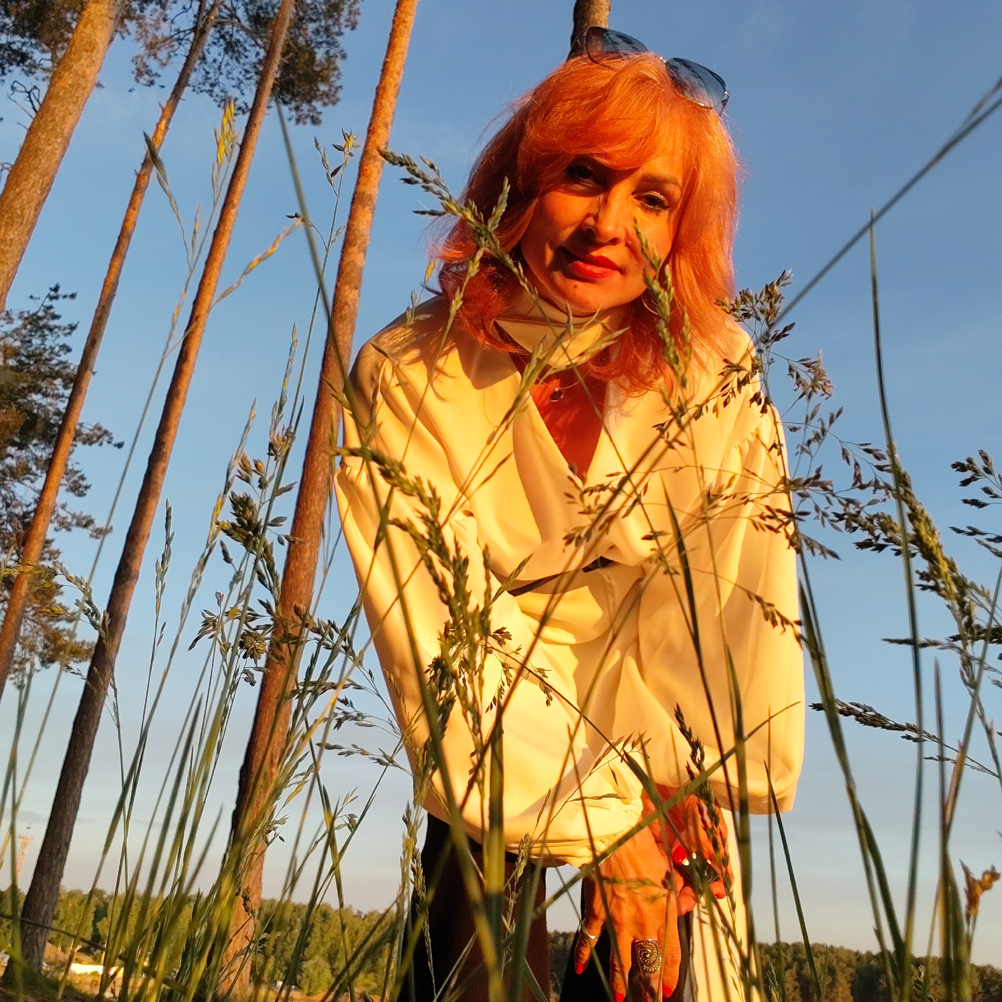 A woman with reddish-orange hair is captured in a low-angle shot, looking towards the camera amidst tall grasses. She is wearing a light-colored, possibly cream or light yellow, long-sleeved top and has sunglasses perched on her head. Her nails are painted red, and she wears a large ring on one finger. The scene is outdoors in a natural setting, likely a grassy field or clearing, with tall pine trees visible in the background against a clear blue sky. The warm, golden light suggests it is golden hour, casting a glow on her hair and clothing and highlighting the tree trunks. The prominent grass in the foreground creates a soft, natural frame for the woman. The overall impression is a serene outdoor portrait. The location is Chaikovsky, Russia. There is no visible text.