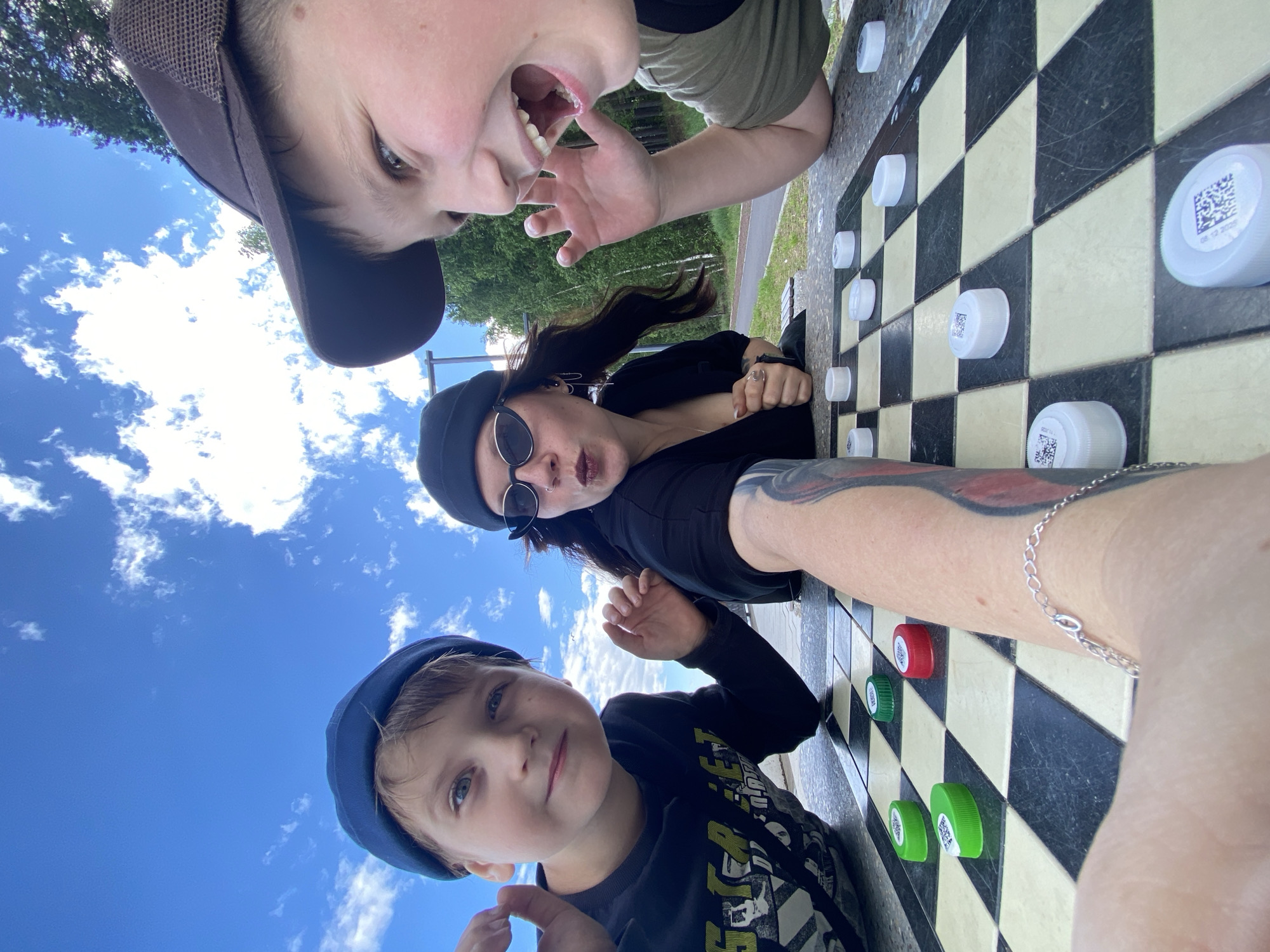 The image depicts a group of three people, two children and one adult, playing checkers outdoors. The checkers board is set on a stone table. The people appear to be engaged in the game, with one child looking directly at the camera and smiling widely, while the other child and the adult are focused on the board. The background shows trees and a blue sky with clouds, suggesting a park or recreational area. The location is identified as the city of Chaikovsky, Russia.