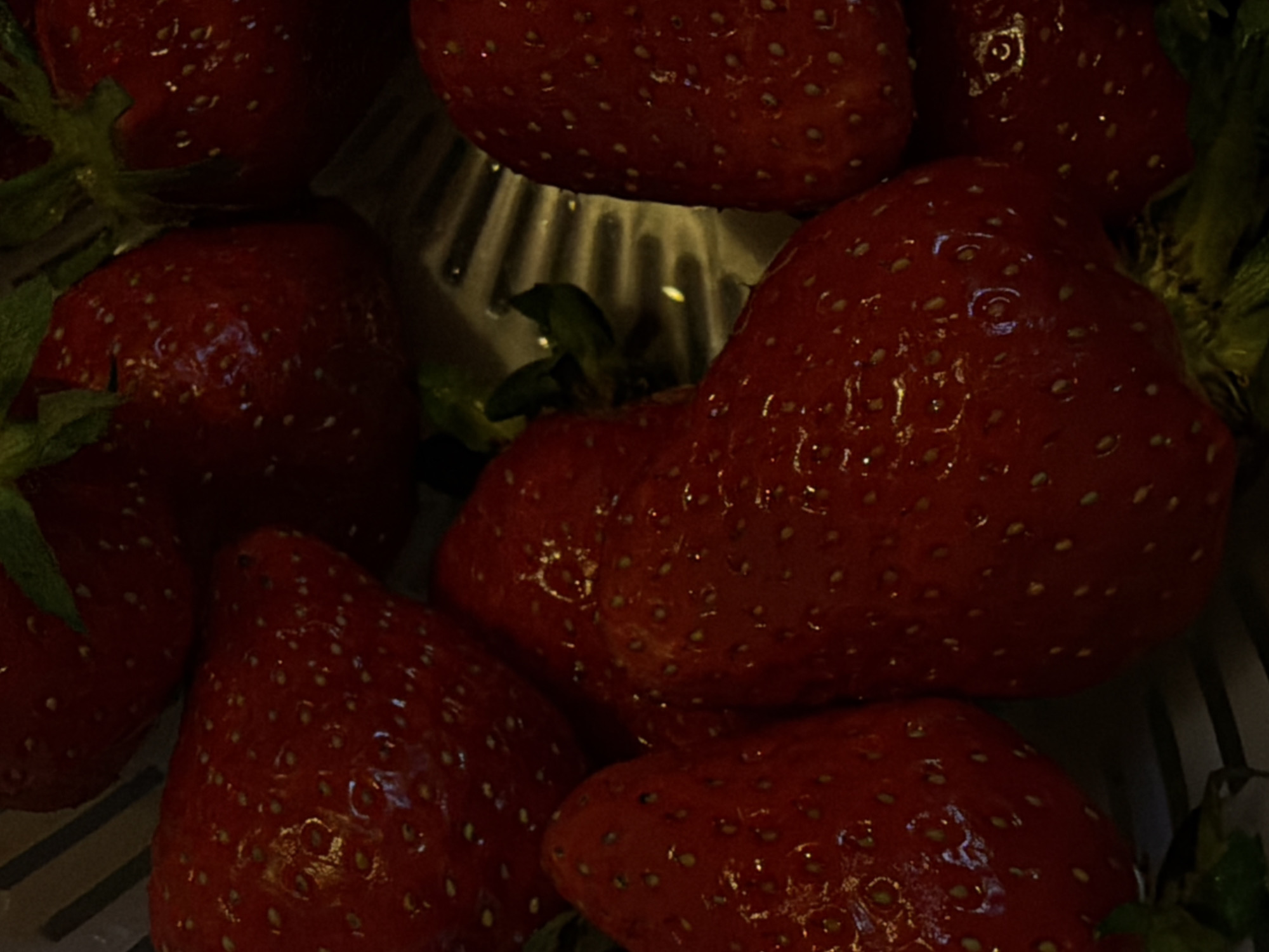 A close-up, dimly lit photograph captures a pile of fresh, ripe strawberries. The primary subjects are several plump, deep red strawberries, many of which still have their vibrant green calyxes (caps) and stems attached. Water droplets are visible on the surface of the fruit, indicating they have either been recently washed or are wet from dew.
The strawberries are arranged in what appears to be a colander or a similar slotted container, with the patterned lines of the base visible through the gaps between the fruit. The dark background and the way light reflects off the wet surfaces suggest an indoor setting, possibly under artificial light or in a low-light environment. The focus is sharp on the strawberries, highlighting their bumpy texture and the tiny seeds embedded in their skin. There is no visible human activity, but the presence of washed fruit implies preparation for consumption. No text is visible in the image. The overall impression is one of freshness and rich, natural color.