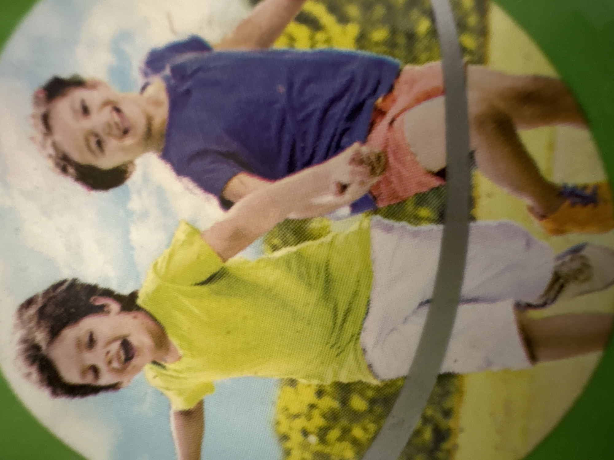 Two young children are depicted running outdoors, holding hands. The child on the left wears a bright yellow t-shirt and white shorts, while the child on the right is dressed in a dark blue t-shirt and coral-colored shorts. They are positioned in front of a blurred background of greenery and a pale blue sky with clouds. A gray horizontal band crosses the image in front of the children. The overall scene suggests a playful, active moment, possibly a race or a game. The image lacks any specific landmarks or identifying features to place it in Ashvem, India.