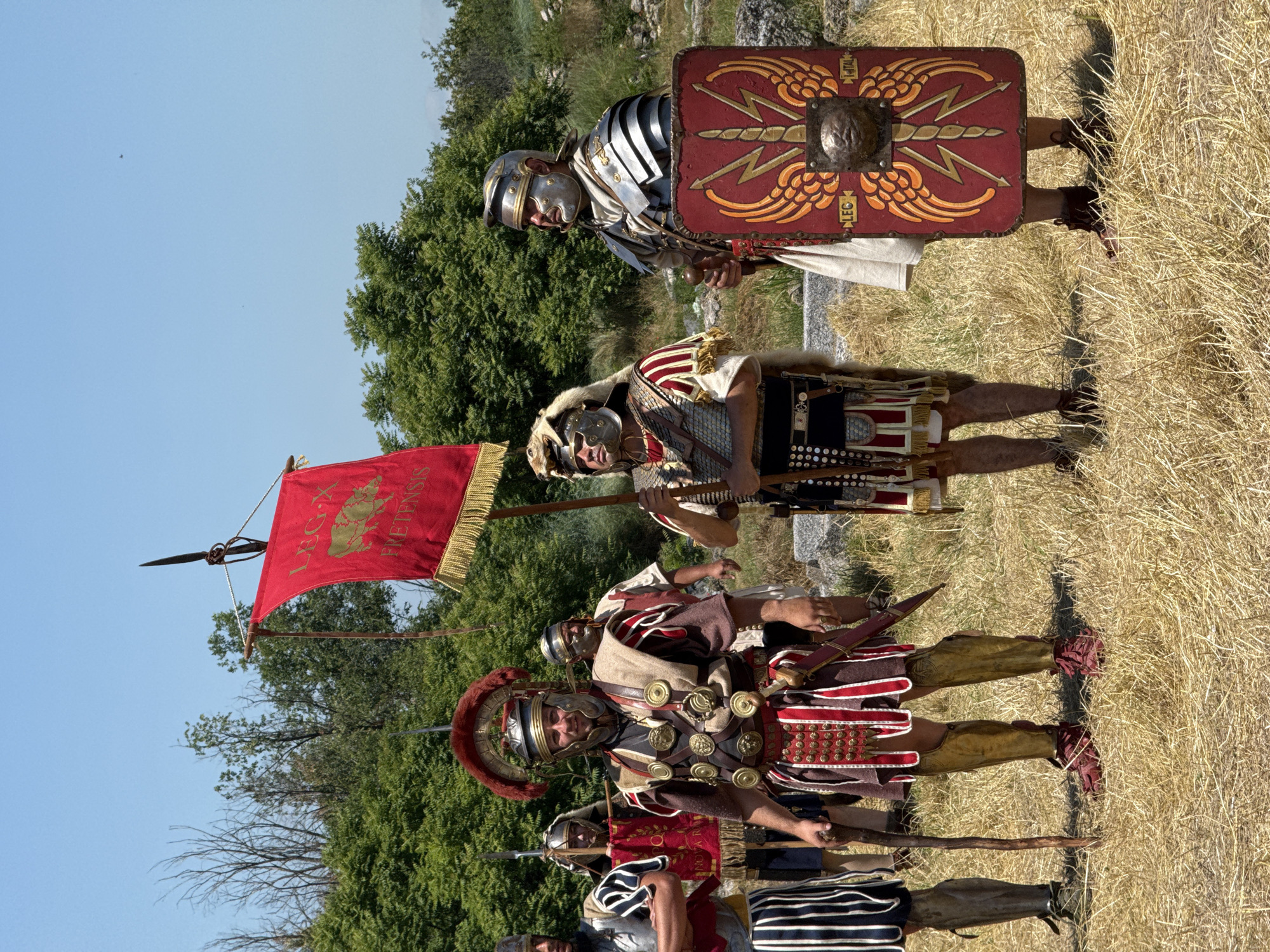 The image depicts an outdoor scene in Gigen, Bulgaria, under a clear blue sky. Six adult males, costumed as Roman soldiers, stand on dry, yellowish grass. In the background, dense green foliage and scattered light-colored stones, suggestive of ruins, are visible.
From left to right:
1. A male in a striped tunic and silver-colored arm protection is partially visible.
2. A central figure wears a plumed helmet, elaborate chest armor adorned with multiple bronze medallions (phalerae), red and white striped pteruges, and golden greaves. A gladius is sheathed at his right hip. He holds a gnarled wooden staff in his left hand and faces slightly right.
3. Behind and to the left of the central figure, another man in a helmet holds a red banner partially obscured, displaying "NOVA ROMA".
4. To the right of the central figure, a standard-bearer (signifer) wears a helmet covered with an animal pelt, chainmail armor (lorica hamata), and a red and white tunic. He holds aloft a red vexillum with golden fringe, depicting a boar and inscribed "LEG X FRETENSIS" in gold lettering. The vexillum is topped with a spearhead finial.
5. To the right of the standard-bearer, a soldier in a silver-colored segmented armor (lorica segmentata), a helmet, and a cream-colored tunic holds a red rectangular scutum (shield). The shield is emblazoned with a golden winged thunderbolt design and a central boss featuring a human or animal face. Inscriptions "LEG" and "I ITAL" are visible on the shield.
6. The rightmost figure is partially visible, wearing a helmet with an animal pelt and chainmail armor, similar to the standard-bearer.
All individuals are standing upright, facing generally towards the viewer or slightly angled. The lighting suggests a sunny day.