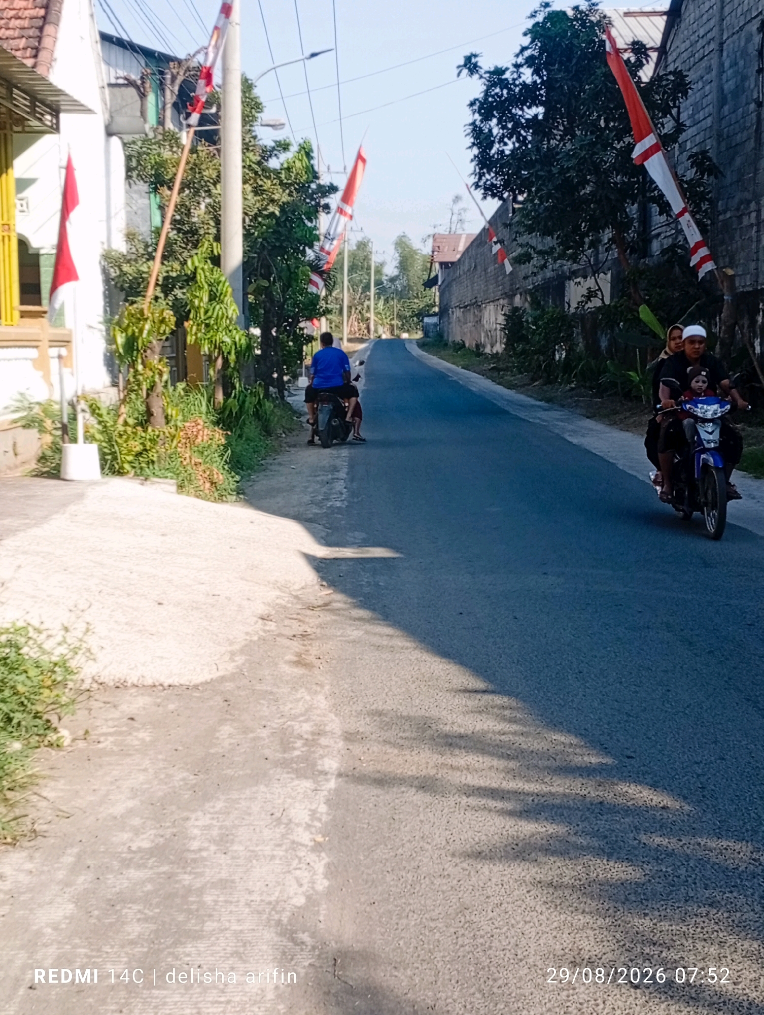 The image depicts a street scene in Coban Blimbing, Indonesia. The road is paved and appears to be a two-lane street with one lane visible. On the right side of the road, a man, a woman, and a child are riding a motorcycle. The man is driving, the woman is behind him, and the child is in front of the man. They are all looking forward. On the left side of the road, a man in a blue shirt is also riding a motorcycle. The environment is characterized by buildings on either side of the road, including a white building with a yellow trim on the left and a gray brick building on the right. Several trees are present along the road. Indonesian flags, red and white, are prominently displayed on poles along the street, suggesting a national holiday or celebration. The lighting suggests it is daytime, with shadows cast by trees on the road. A timestamp in the lower right corner reads "29/08/2026 07:52". A watermark in the lower left corner reads "REDMI 14C | delisha arifin".