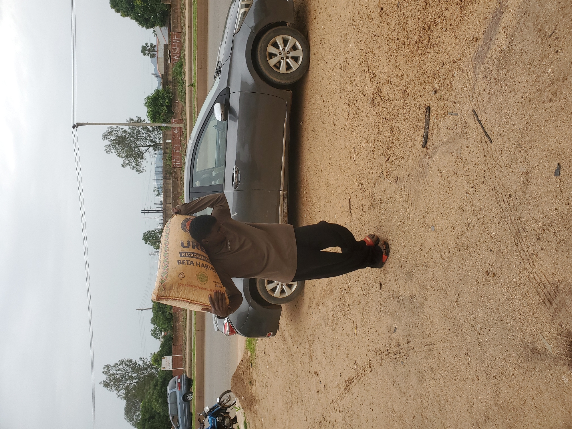 A man carries a large sack of grain over his shoulder, walking on a dusty path. He is wearing a brown long-sleeved shirt and black pants. He has red sandals on his feet. A gray sedan is parked behind him on a paved road. In the background, there are trees, a blue minivan, and utility poles. The sky is overcast. The scene is in an urban area in Unknown, Nigeria.