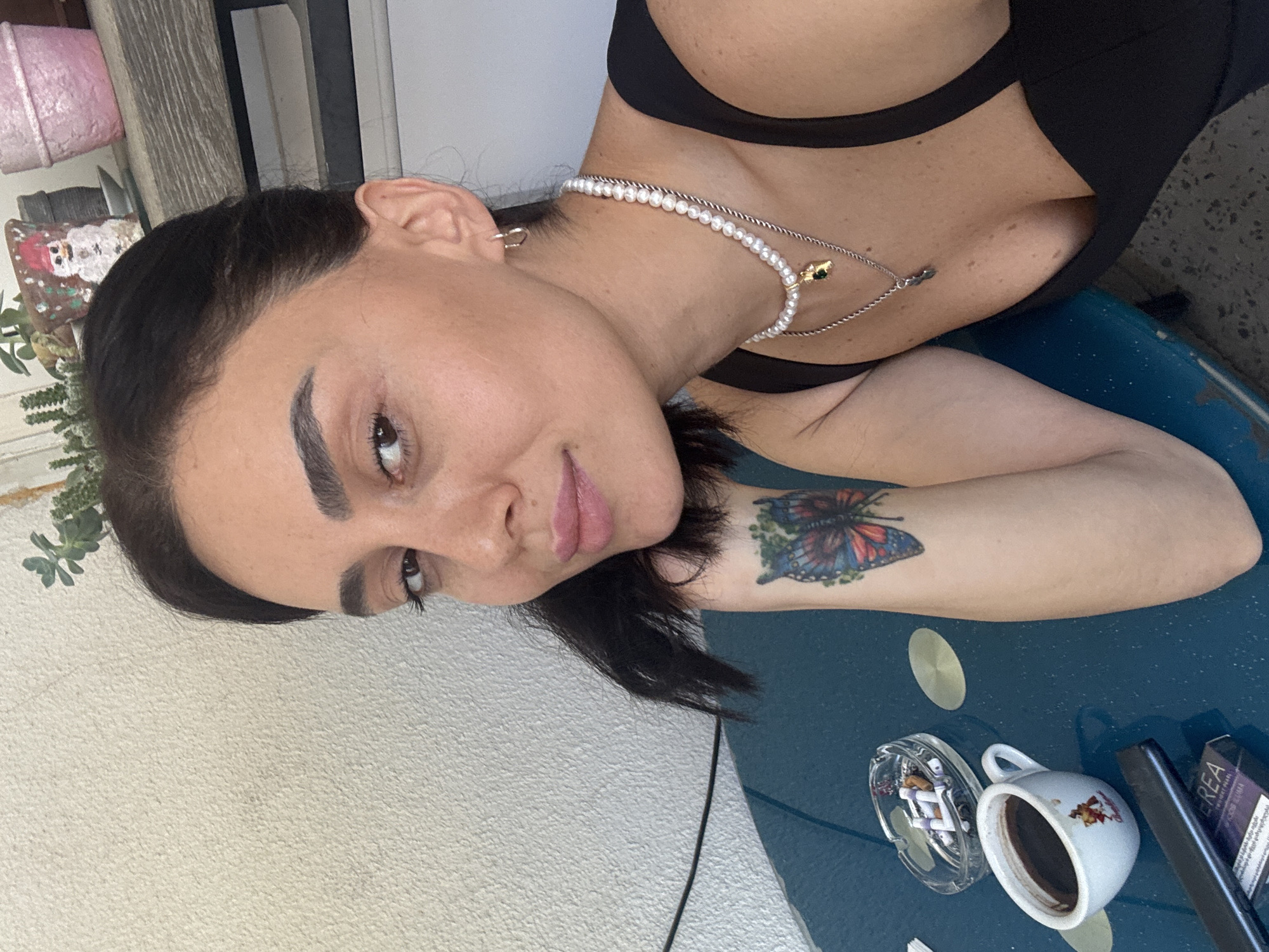 A woman is captured in a close-up shot, her face angled towards the camera with a subtle smile. She has dark hair pulled back, and her makeup emphasizes her eyes and lips. She is wearing a black top with a revealing neckline, adorned with a pearl necklace and a silver pendant. On her left forearm, a colorful butterfly tattoo is visible. In the foreground, a blue table holds a coffee cup, an ashtray with cigarette butts, and a pack of cigarettes. The background is a plain, textured wall with a shelf above, featuring decorative pots and a plant. This scene appears to be an outdoor setting, possibly a balcony or patio, in Tbilisi, Georgia, given the context.