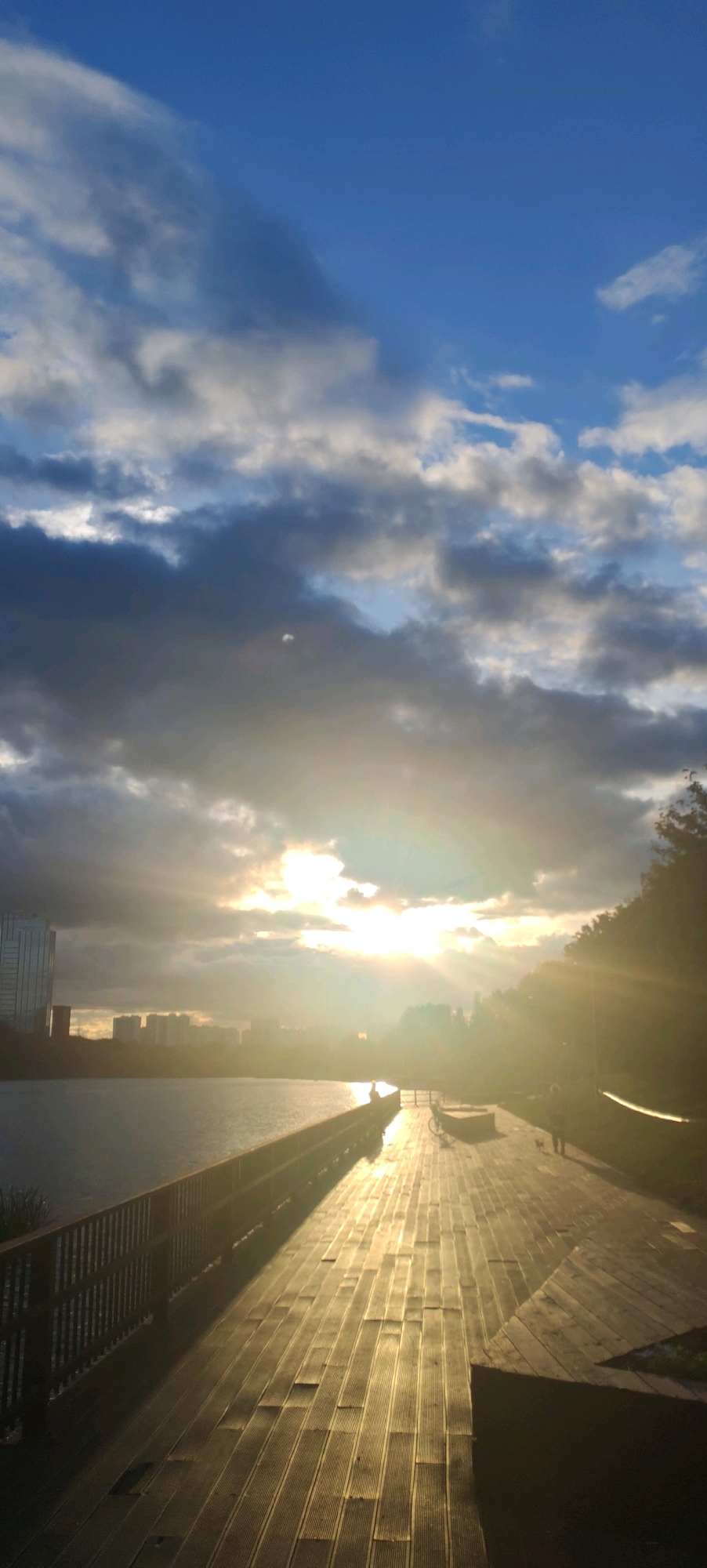 This is a photograph of a waterfront promenade at sunset, likely in Moscow, Russia. The sun is low in the sky, casting long shadows and creating a warm, golden light that reflects off the water and the wooden planks of the promenade. The sky is a mix of blue and dramatic grey clouds, with the sun breaking through in a bright, hazy glow.
In the foreground, the wooden promenade stretches into the distance, with a wooden railing along the left side separating it from the water. The planks are oriented perpendicular to the direction of travel, with visible lines and slight gaps between them.
On the left side of the image, a modern glass skyscraper stands tall, suggesting an urban environment. Across the water, a city skyline with buildings of varying heights is visible in the distance, partially obscured by haze and the setting sun.
Several figures are present on the promenade. To the right of the center, a silhouette of a person is visible near a bicycle. Further along the promenade, another person is seen walking with a small dog. The atmosphere appears to be calm and serene, typical of an evening stroll.
The location is consistent with the Moscow River embankment or a park area with a body of water and walkways, common in areas such as Luzhniki Park or areas along the Moscow Canal. The presence of modern buildings and the general landscape align with urban development found in Moscow.
