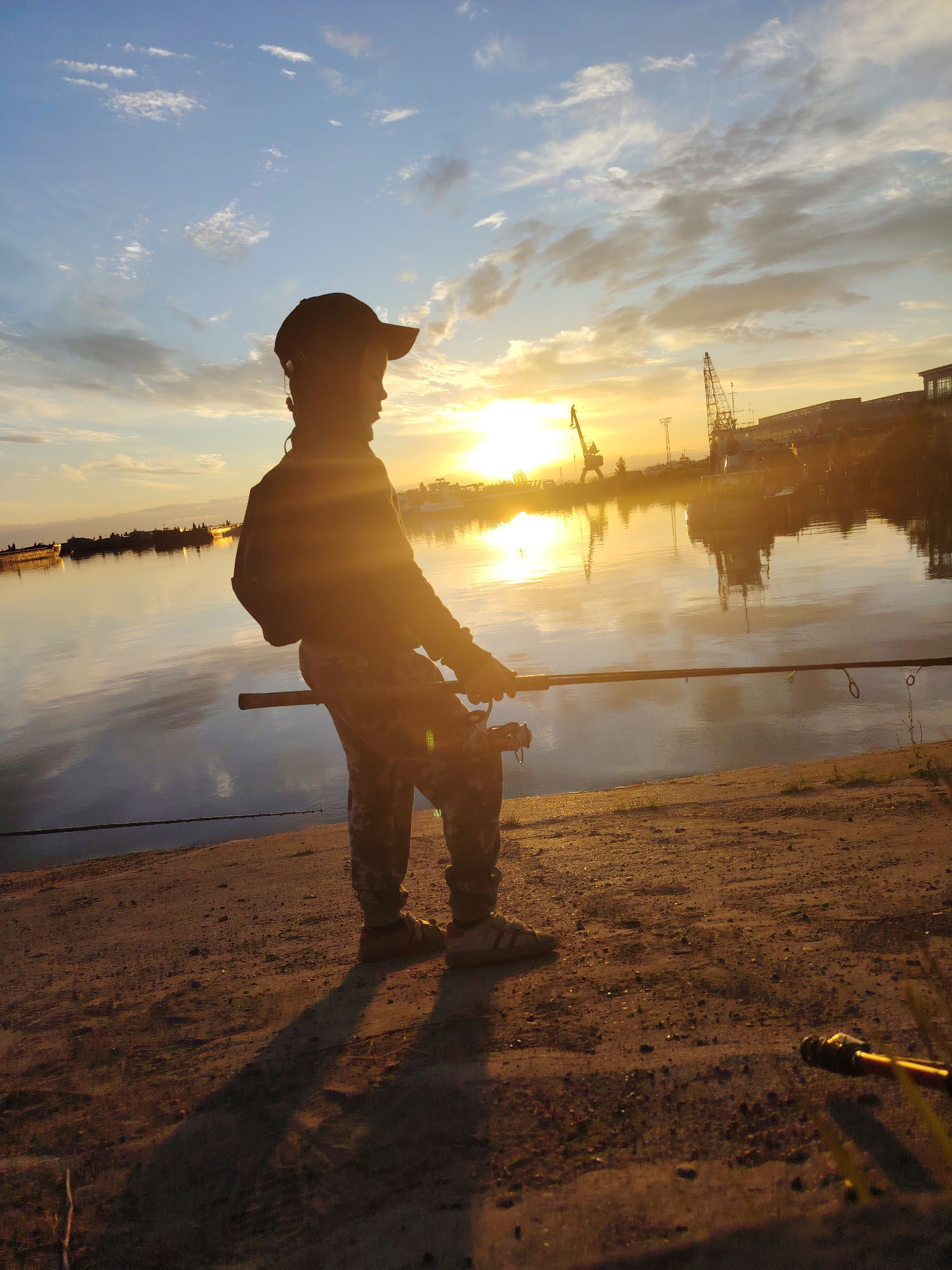 A young boy, silhouetted against a setting sun, stands on the muddy bank of the Kama River in Chaikovsky, Russia, holding a fishing rod. The boy wears a baseball cap, a dark jacket, and camouflage pants. His shadow stretches long across the ground. In the distance, industrial cranes and ships are visible across the water, reflecting in the calm surface of the river. The sky is a mix of blue and orange hues, with scattered clouds. The overall scene evokes a peaceful, end-of-day atmosphere.