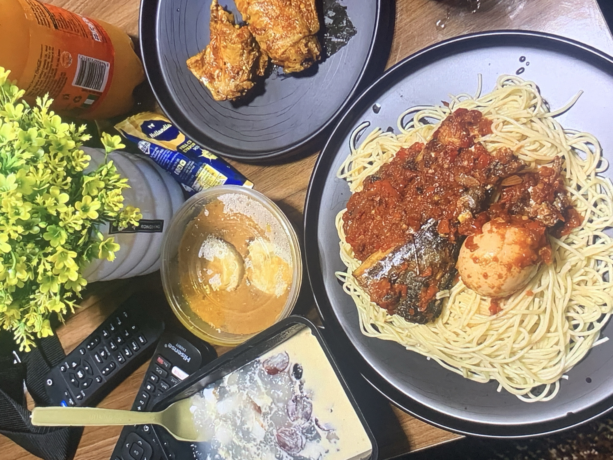 The scene depicts a tabletop still life in Unknown, Unknown, featuring various food items and other objects. A black plate on the upper left contains what appears to be fried food. Another black plate on the right holds spaghetti with tomato-based sauce, pieces of fish, and a hard-boiled egg. A clear container filled with a beige-colored substance and slices of banana is present. A dark rectangular container with a lid ajar contains a light-colored substance mixed with what appear to be dark fruits. An orange-colored bottle is positioned on the upper left, along with a package of Hollandia yogurt, a small potted plant with yellow flowers, and two television remote controls. The spoon rests atop one of the remotes. The table surface is wooden.