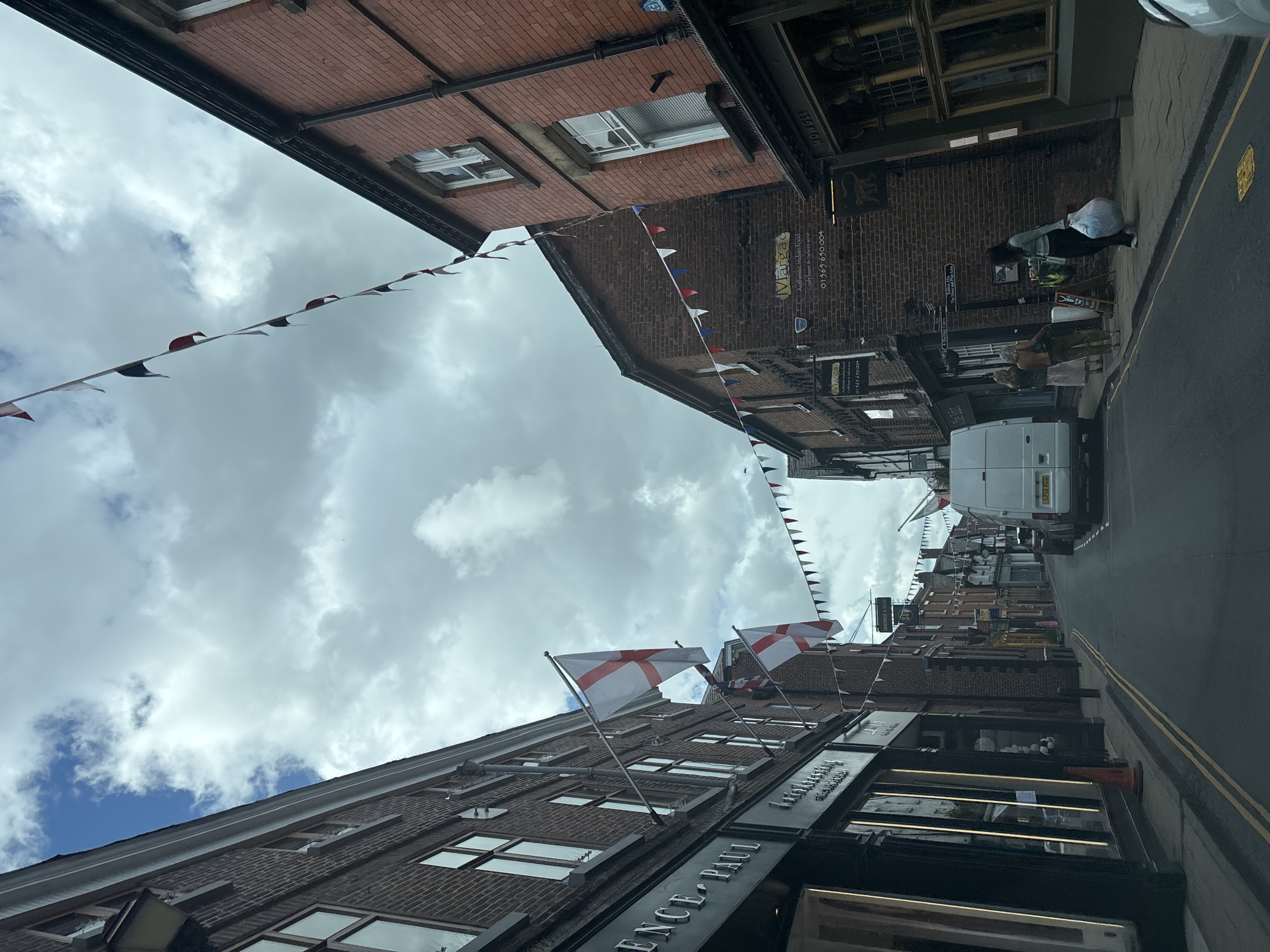 Here is a description of the image:
The image shows a street in Knutsford, United Kingdom, with a slightly tilted perspective. Buildings line both sides of the street, primarily constructed of red brick. Festive bunting and English flags are strung across the street, adding a celebratory atmosphere. A white van is parked on the street. People are seen walking on the sidewalk. Shop signs indicate businesses such as "Clarence Paul" and "Margao" restaurant. The sky is cloudy. Double yellow lines are marked on the road. The photograph appears to be taken from a low angle within the street itself.