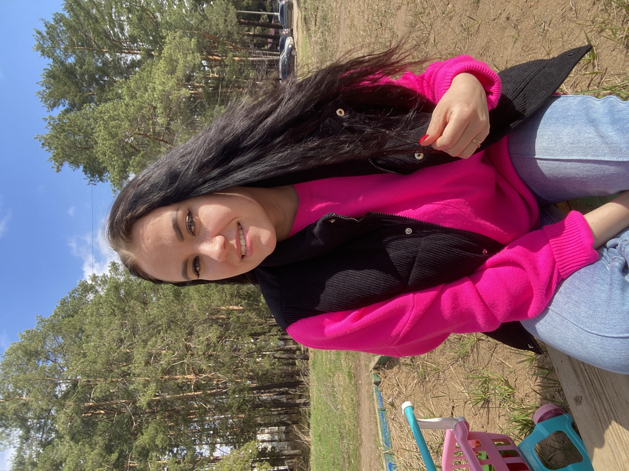 This image, taken in Chaikovsky, Russia, captures a young woman smiling and looking directly at the camera while outdoors. She has long, dark hair and is wearing a vibrant fuchsia pink long-sleeved sweater underneath a black vest or jacket, paired with light blue jeans. Her left hand is visible, adorned with red nail polish and a ring on her ring finger.
The setting appears to be a natural recreational area or park, with a backdrop dominated by tall, green coniferous trees, likely pines, under a clear blue sky dotted with a few white clouds. The ground is dry, covered with sparse grass and patches of bare earth, hinting at early spring or a dry season. In the distance, among the trees, several parked cars can be seen.
In the foreground, toward the bottom right, there are brightly colored children's toys: a small, light blue and pink plastic toy cart or stroller, and another light blue and pink plastic toy, possibly a chair. The woman's relaxed pose and the presence of children's toys suggest she is enjoying a leisurely day outdoors, perhaps with a child who is not in the frame. The scene is brightly lit by natural sunlight. No visible text is present in the image.