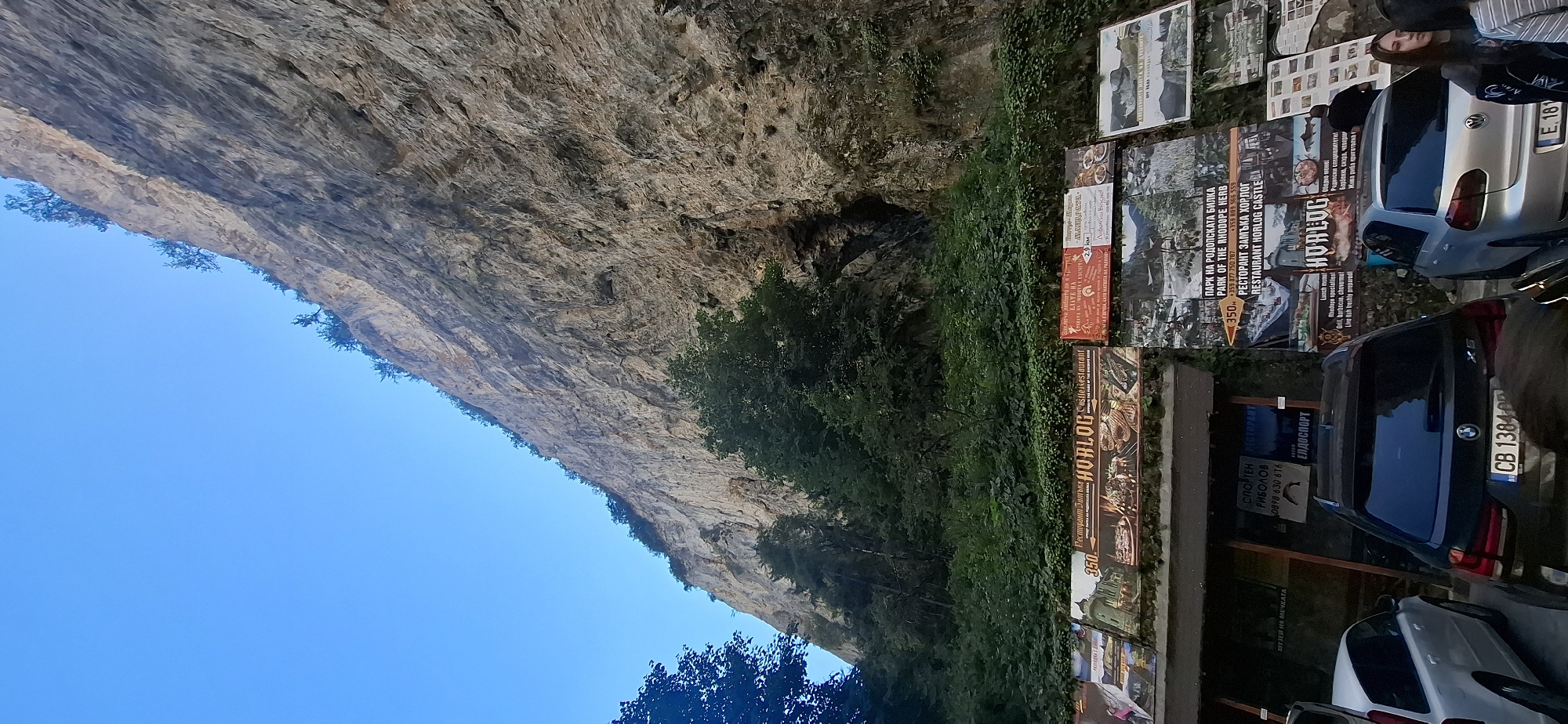 The image depicts a scenic view of a towering cliff face next to a road with parked cars in Trigrad, Bulgaria. The cliff, predominantly grey and rocky, features sparse vegetation clinging to its upper edges and a small cave-like opening partway up. At the base of the cliff, a row of advertisements is affixed to a covered structure, likely a restaurant or tourist information center. These advertisements display images of food, landscapes, and text in what appears to be Bulgarian. One prominent sign reads "Horlog Castle Restaurant." Several cars are parked in front of the structure, including a silver BMW SUV with Bulgarian license plate CB 1384 OP, a white Volvo station wagon, and a silver Volkswagen hatchback. A few people are visible in the foreground, near the parked cars. The sky is clear and blue. The overall impression is a tourist destination nestled in a dramatic natural landscape.