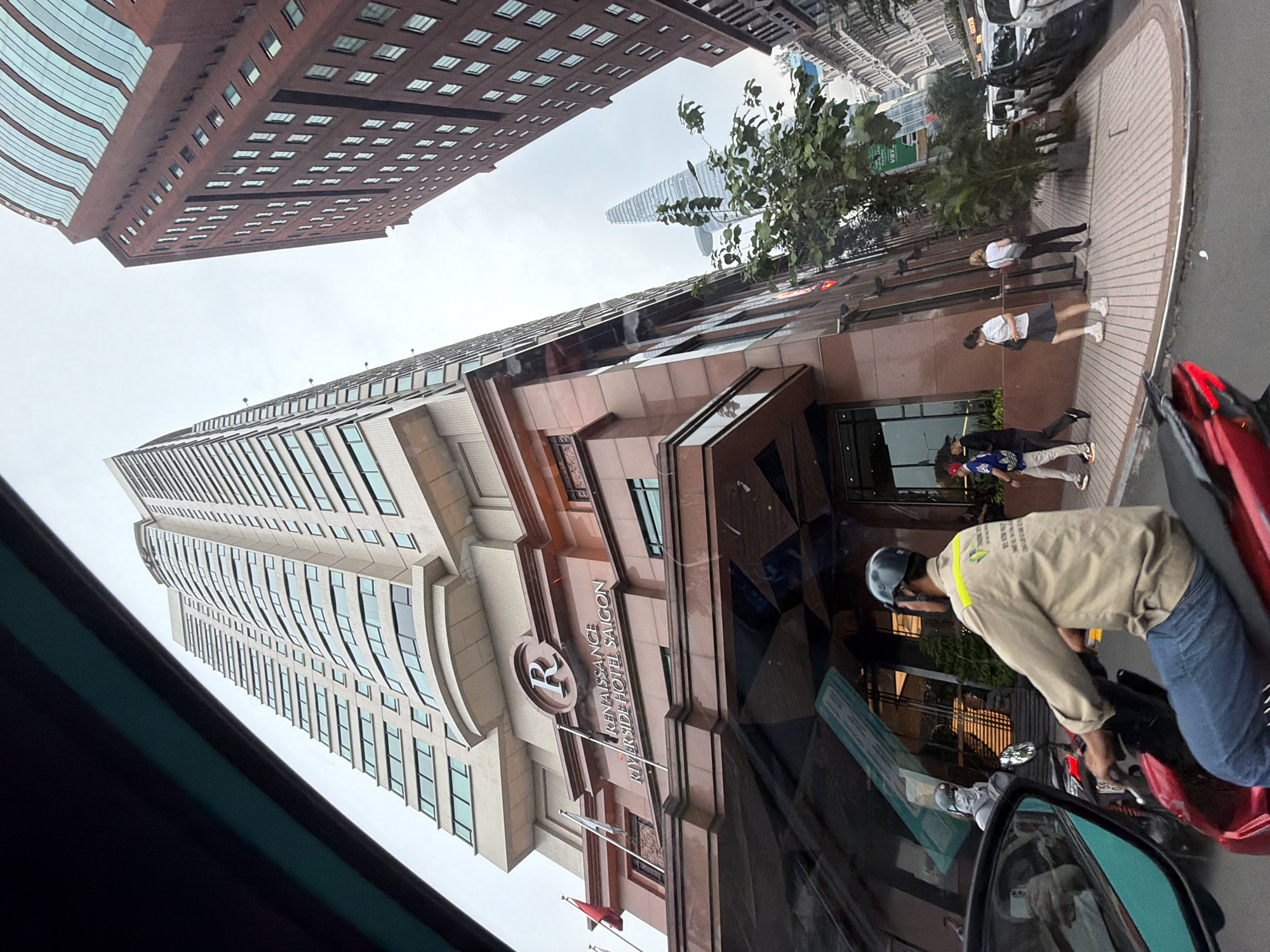 The image shows the exterior of the Renaissance Riverside Hotel Saigon, a tall building with a reddish-brown facade. The hotel's name and logo are prominently displayed on the building. In the foreground, a person is riding a red scooter, wearing a beige uniform and a helmet. Several other people are walking on the sidewalk in front of the hotel. The sky is overcast, and other tall buildings are visible in the background, characteristic of a city environment. The location is in the city of Thủ Đức, Vietnam.