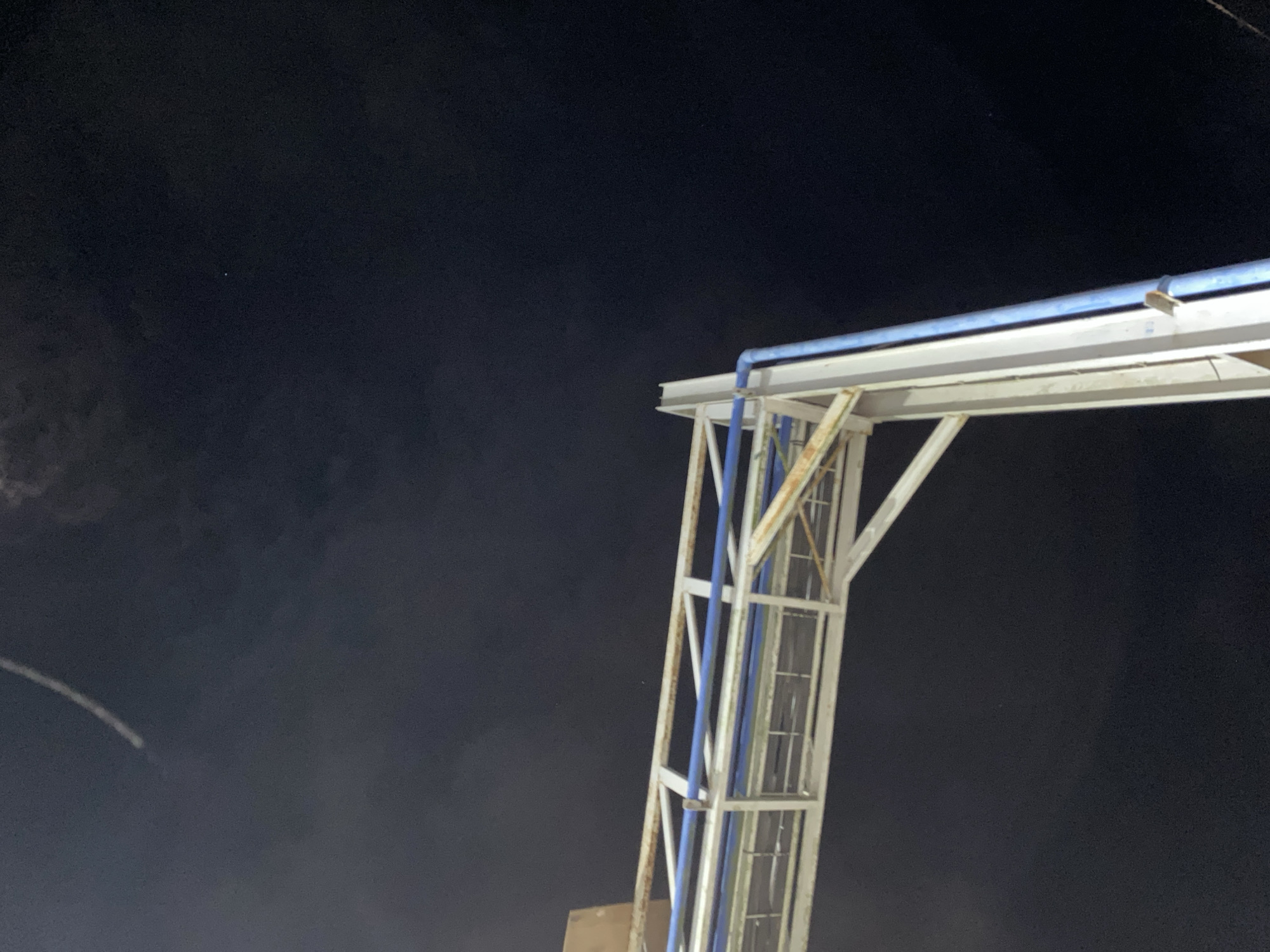 The image shows a nighttime view looking up at a metal structure, possibly part of an overpass or a support for utilities, against a dark sky. The structure is white with some rust showing and has blue pipes running along it. In the upper left quadrant of the sky, there is a faint white streak, possibly a contrail from an aircraft or a shooting star. A single small bright point, likely a star or planet, is visible in the upper right quadrant of the sky. The background is a deep, dark blue, indicating it is nighttime. The scene appears to be in an outdoor urban or semi-urban environment. There are no people or animals visible. The lighting is dim, suggesting artificial light sources are not prominently illuminating the sky.