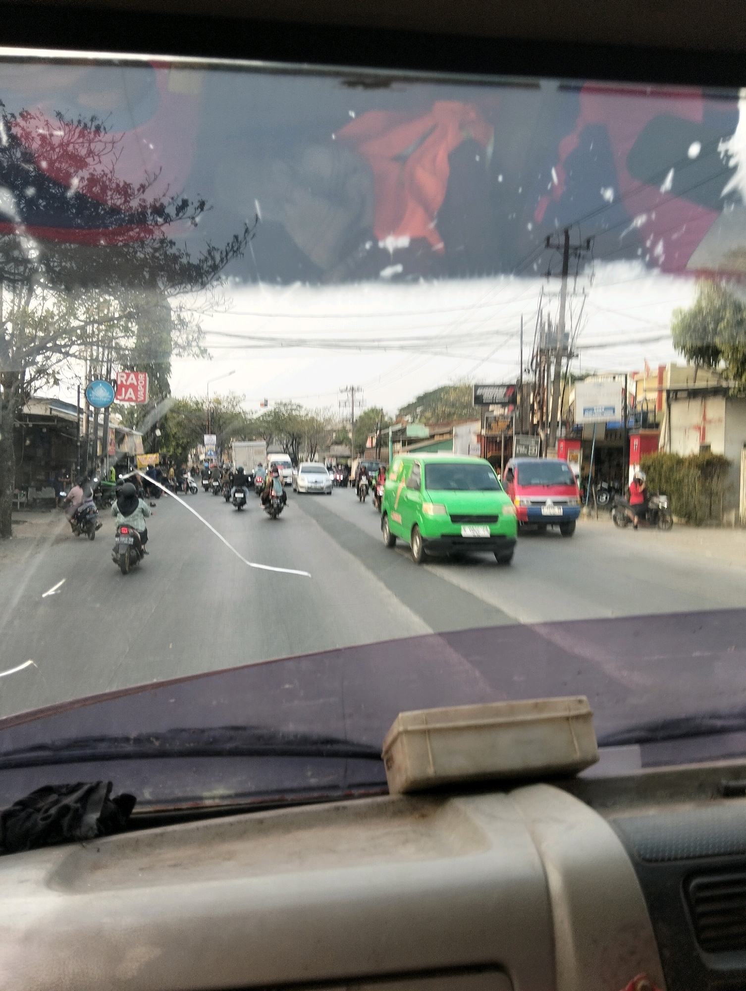 The image captures a view from inside a vehicle, looking out through the windshield onto a street in Semarang, Indonesia. The sky is overcast. In the foreground, the dashboard of the vehicle is visible, with a grey rectangular object placed on it. The windshield shows reflections of trees and possibly a building with red elements.
The street scene itself is busy with traffic. Several motorcycles are in motion, with riders wearing helmets and casual clothing. A green van is prominently positioned in the middle of the road, followed by a red van. Other vehicles, including cars and trucks, are also present in the traffic flow. Buildings line the street, some appearing to be shops or businesses with signage. Power lines and utility poles are visible in the background, typical of an urban environment. The road surface is asphalt, with lane markings. The overall impression is of a moderately busy street in a city.