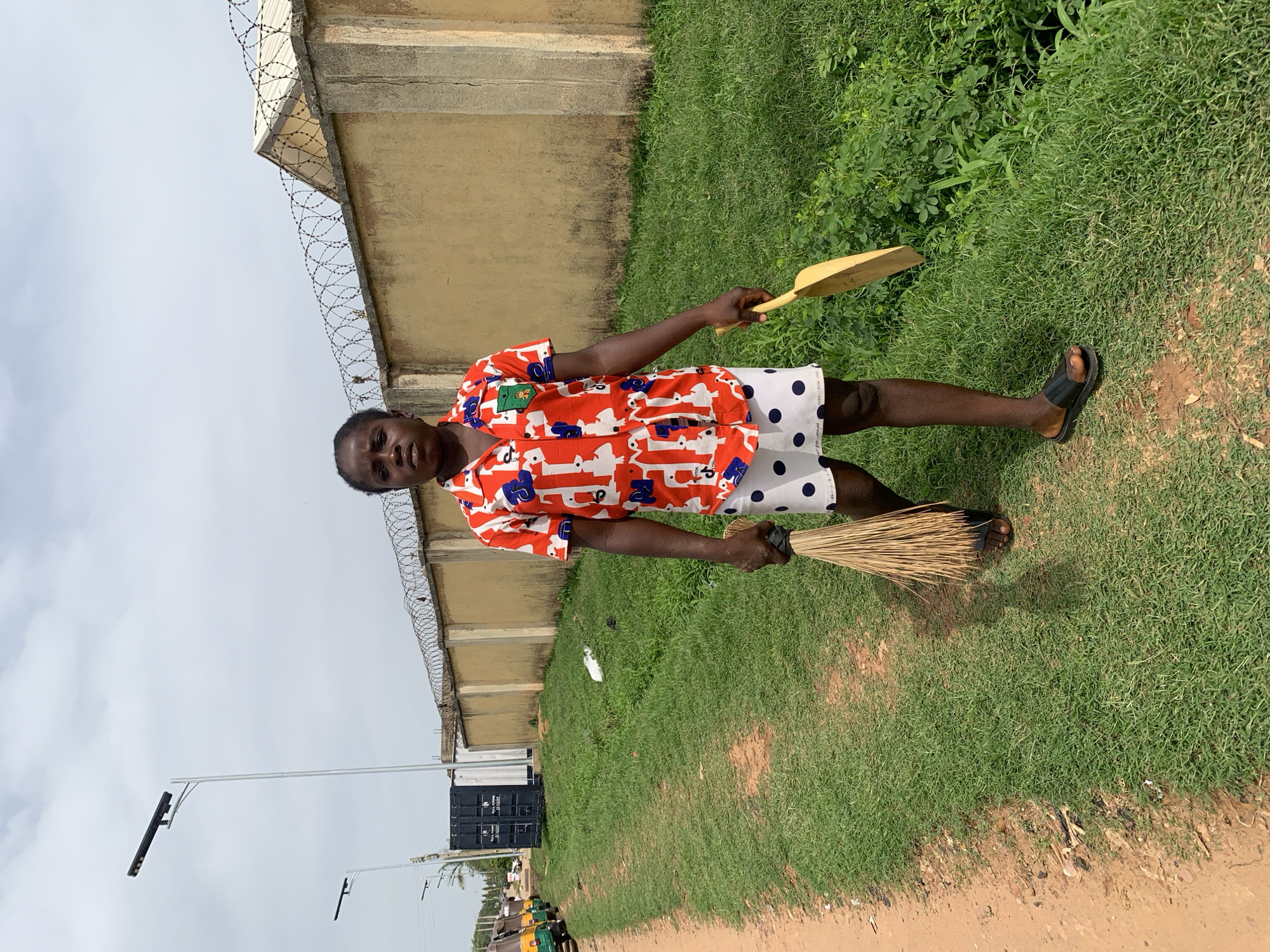 A young Black individual with short hair, wearing a red short-sleeved shirt with white abstract patterns and a white skirt with navy blue polka dots, stands on a dirt path next to a grassy area. They are holding a straw broom in their left hand and a yellow dustpan in their right hand. The person is facing the camera and appears to be walking forward. In the background, there is a long, yellowish-beige wall topped with barbed wire, and beyond that, a cloudy sky. To the left of the wall, there are two tall lampposts. Further left, a dark blue shipping container and a line of three yellow and green auto-rickshaws are visible. The overall scene suggests an outdoor environment in a city, possibly on a street or pathway in Jos, Nigeria, given the context provided.