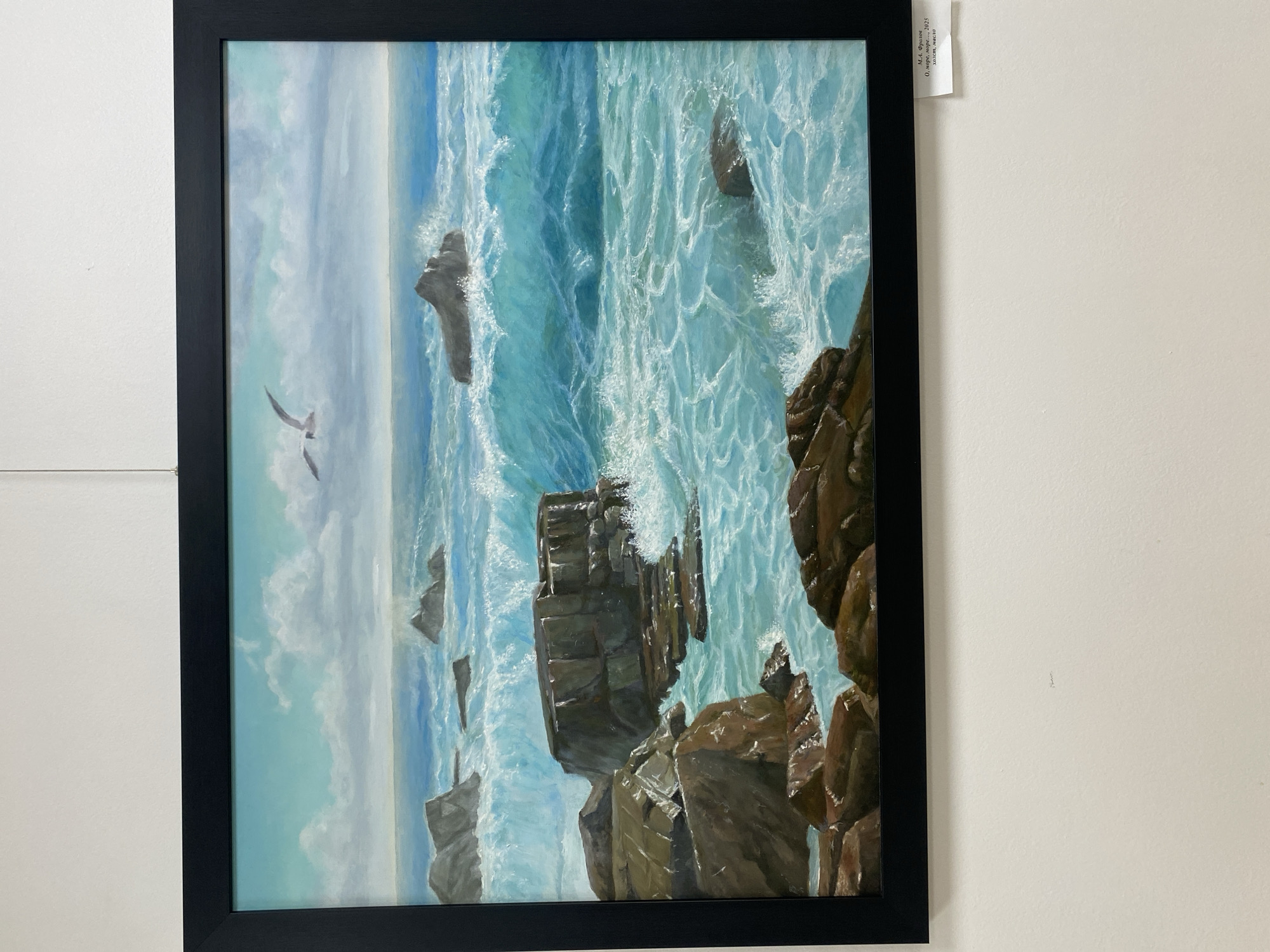 This is a painting of a turbulent sea crashing against rocky shores. A seagull is in flight in the upper portion of the sky, which is filled with clouds. The waves are depicted with vibrant blues and whites, creating a sense of movement and power. Rocks of various sizes and shapes are scattered throughout the foreground and midground, some submerged in the water and others rising above the waves. The painting is framed in black. A small white label is attached to the wall below the painting, to the right. The text on the label is in Russian and appears to be an artist's name, title, date, and medium: "М.А. Фролов / О, море, море..., 2025 / холст, масло". This translates to: "M.A. Frolov / Oh, sea, sea..., 2025 / canvas, oil". The location of this artwork is not specified in the image.