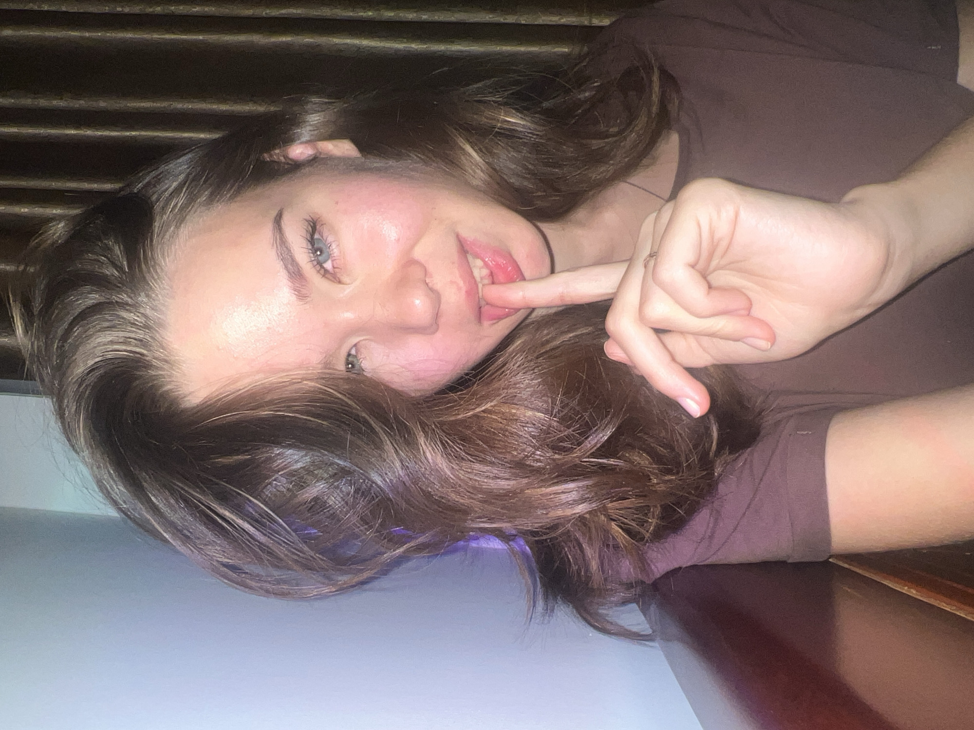 A young woman with long, wavy, dark brown hair is captured in a close-up, horizontal portrait, looking directly at the camera. Her head is tilted, and she appears to be lying down on her side. She has light blue or grey eyes, a soft smile, and her left index finger is gently placed on her lower lip, suggesting a playful or thoughtful expression. She has a natural blush on her cheeks and her lips are glossy. She is wearing a simple dark brown or maroon colored top.
The background behind her head is dark and features horizontal textures, possibly blinds or wooden paneling. Her hair spills over a light blue-grey surface in the foreground, likely a bed or table. To the right, a darker, reddish-brown wooden surface is visible. The lighting is dim, casting a soft, warm glow, indicating an indoor setting, possibly in the evening or at night. There are no discernible location cues within the image that specifically point to Nam Natural Heritage Area Trang, Vietnam; it depicts a generic interior. No text is visible in the image.