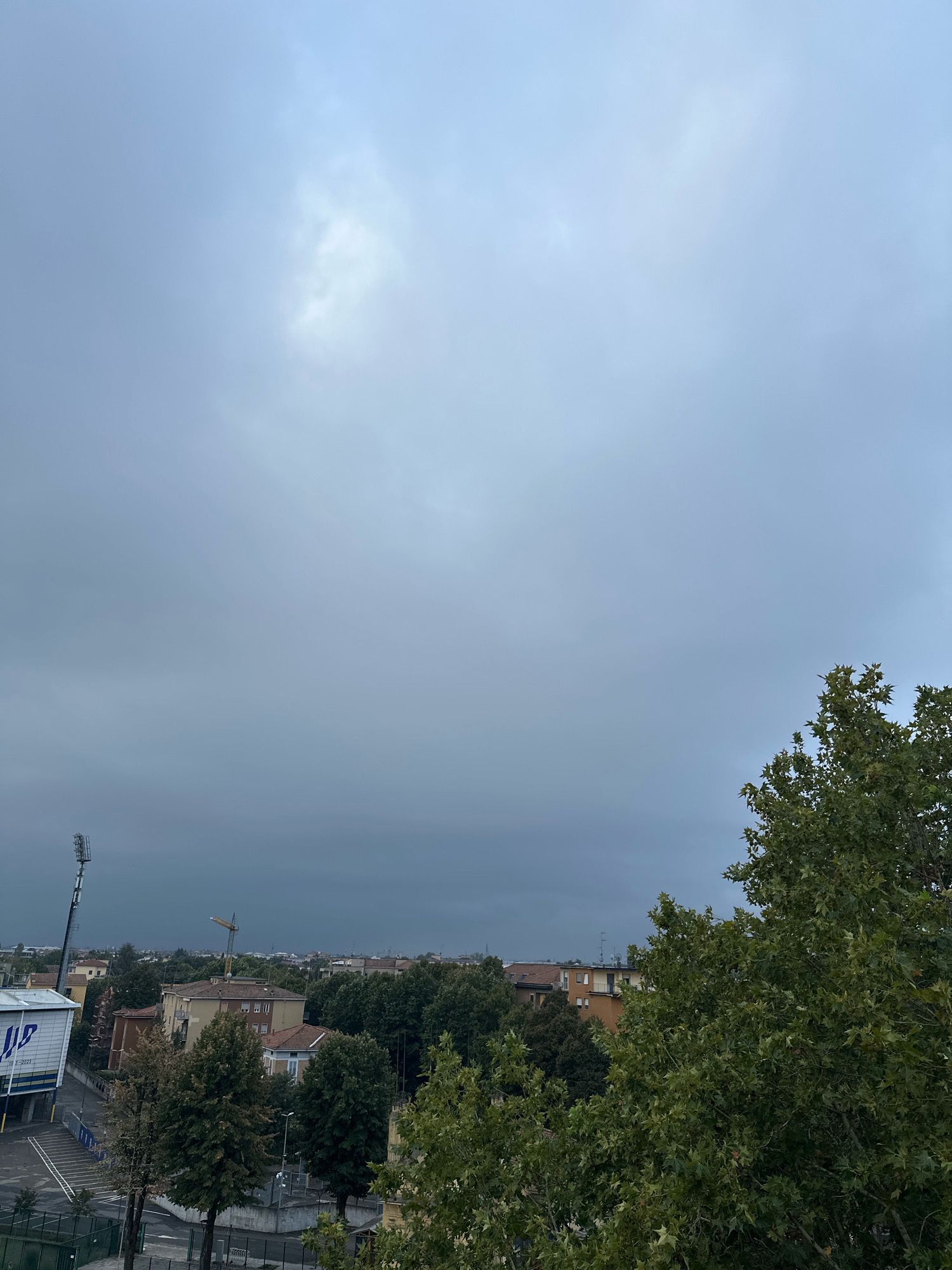 The image captures an overcast sky over a cityscape, with a prominent sports stadium visible on the left. The stadium features a tall lighting tower and a building with blue and white signage. Beyond the stadium, the city unfolds with residential buildings, trees, and a construction crane in the distance. The foreground is partially obscured by the lush green foliage of a large tree. The scene suggests a somewhat somber or pre-storm atmosphere due to the heavy clouds. The city of Parma, Italy is known for its architectural heritage and vibrant cultural scene, often featuring similar urban landscapes with historic buildings interspersed with modern infrastructure and greenery.