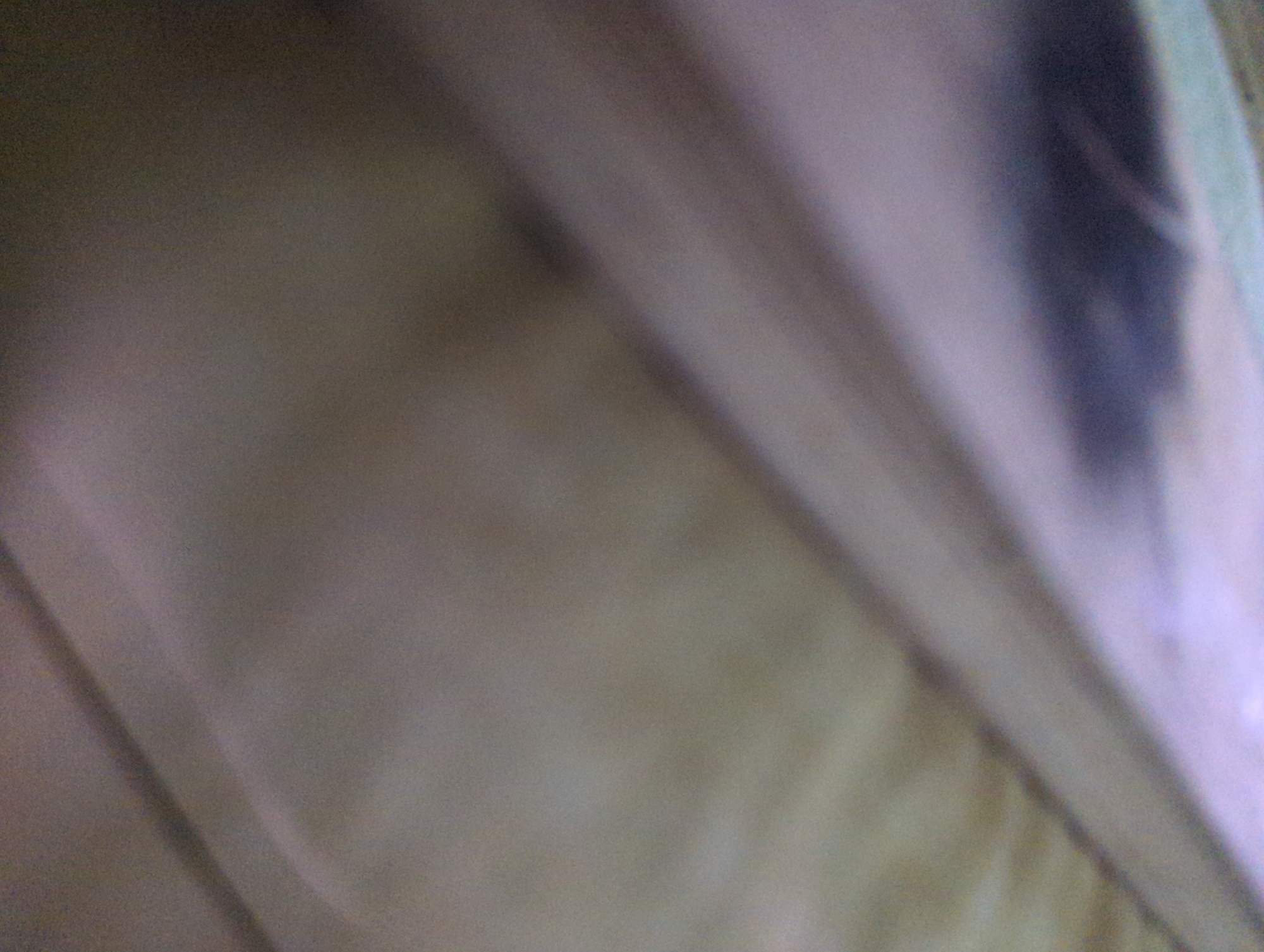The image presents an abstract, heavily blurred, and out-of-focus scene, attributed to the city of Chaguanas, Trinidad and Tobago. The composition consists primarily of muted tones, ranging from light beige-gray to darker purplish-gray and brown. Diagonal lines or folds traverse the frame, originating from the upper left and extending downwards towards the right, creating a sense of layered or textured surfaces. A distinct, amorphous dark gray shape occupies the lower central-right portion of the frame, possibly representing a shadow or a deeply shaded area. The lighting is diffuse, with subtle gradations between lighter and darker regions across the visual field. There are no discernible people, specific objects, or overt actions or interactions depicted. The overall environment appears to be an indistinct, close-up view of a surface, possibly fabric or a wall, under low or uneven illumination.