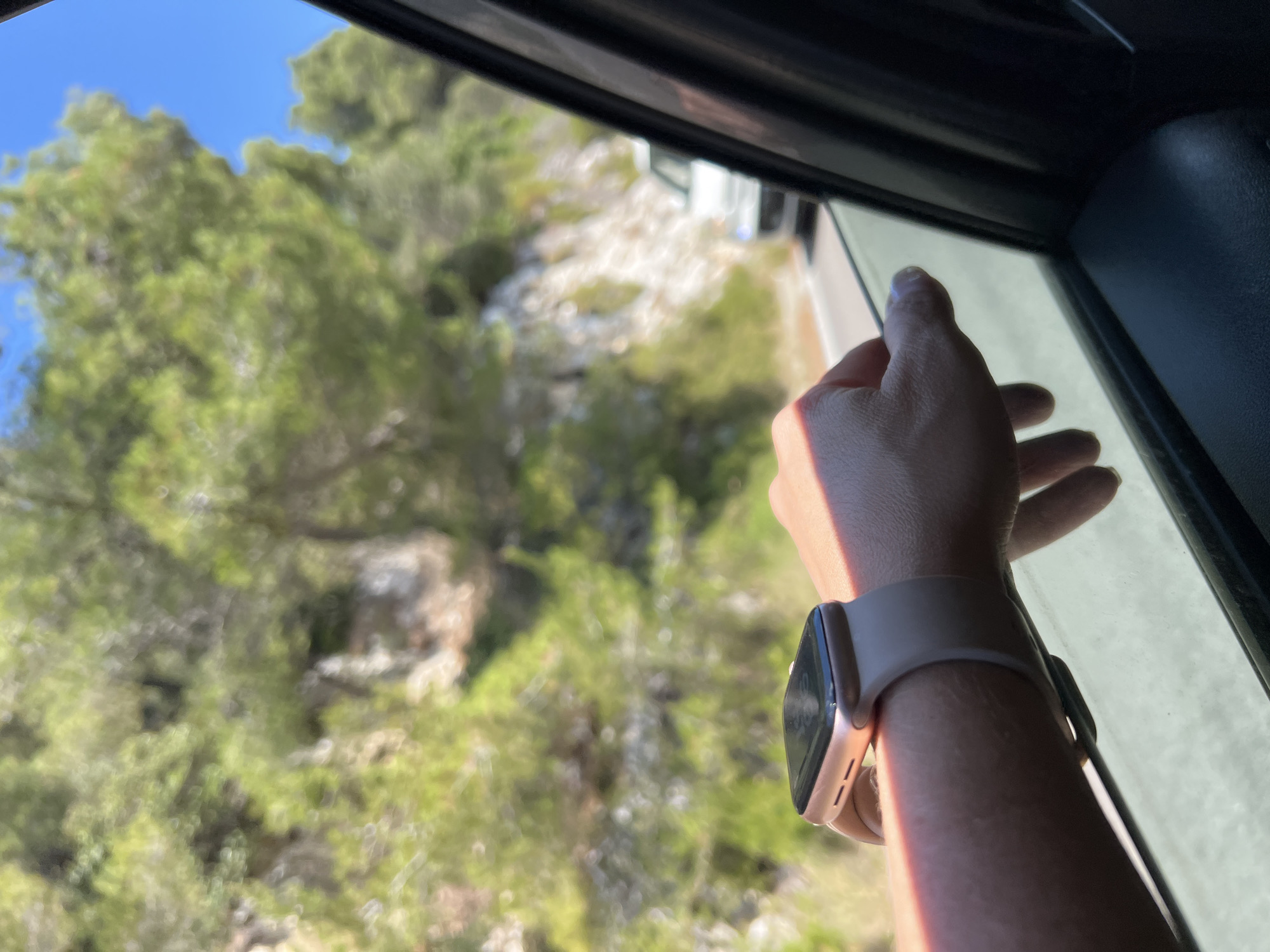 An arm with a smartwatch rests on a car window, with the hand slightly extended outside. The background shows a sunlit coastal road with green foliage and a blue sky. The scene is likely near Mattinata, Italy, a town known for its scenic coastal roads.