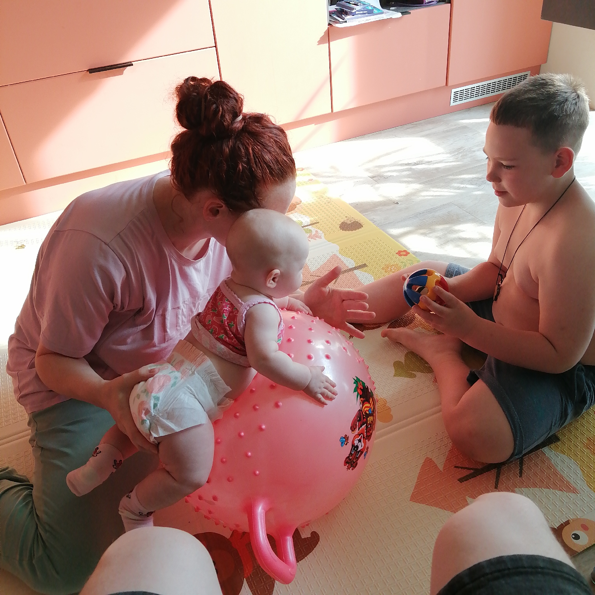 In a room with peach-colored cabinets and a light wood floor, a woman with red hair pulled into a bun kneels, supporting a baby seated on a pink inflatable ball. The baby wears a pink outfit with strawberry print and a diaper. A young boy with short brown hair sits to the right, holding a multi-colored ball. The scene appears to be indoors, possibly in a home setting. The specific location in Trubacheyevka, Russia, is not discernible from the visual information provided.