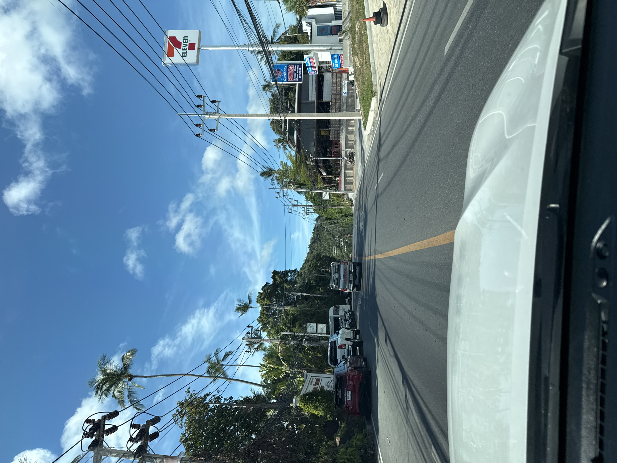 The image depicts a street scene in Baan Chaweng Noi, Thailand, on a clear day with blue skies and scattered clouds. The perspective is from the driver's seat of a vehicle, looking forward down a paved road. On the left side of the road, there are dense green trees and palm trees, a sign that reads "Samui Summit," and several parked vehicles including a red sedan, a white pickup truck, and a white van. Further down the road, a white Toyota pickup truck is visible.
On the right side of the road, a prominent 7-Eleven convenience store sign is visible. Adjacent to it is a building with a roof extending over a dining area, with tables and chairs. A currency exchange sign with blue lettering is also present. A few motorcycles are parked near the buildings, and a person is seen riding a motorcycle. Electrical poles with numerous wires run along both sides of the street. The road is marked with a yellow center line and has shadows cast by overhead wires. A traffic cone made of stacked tires is present on the right shoulder of the road.