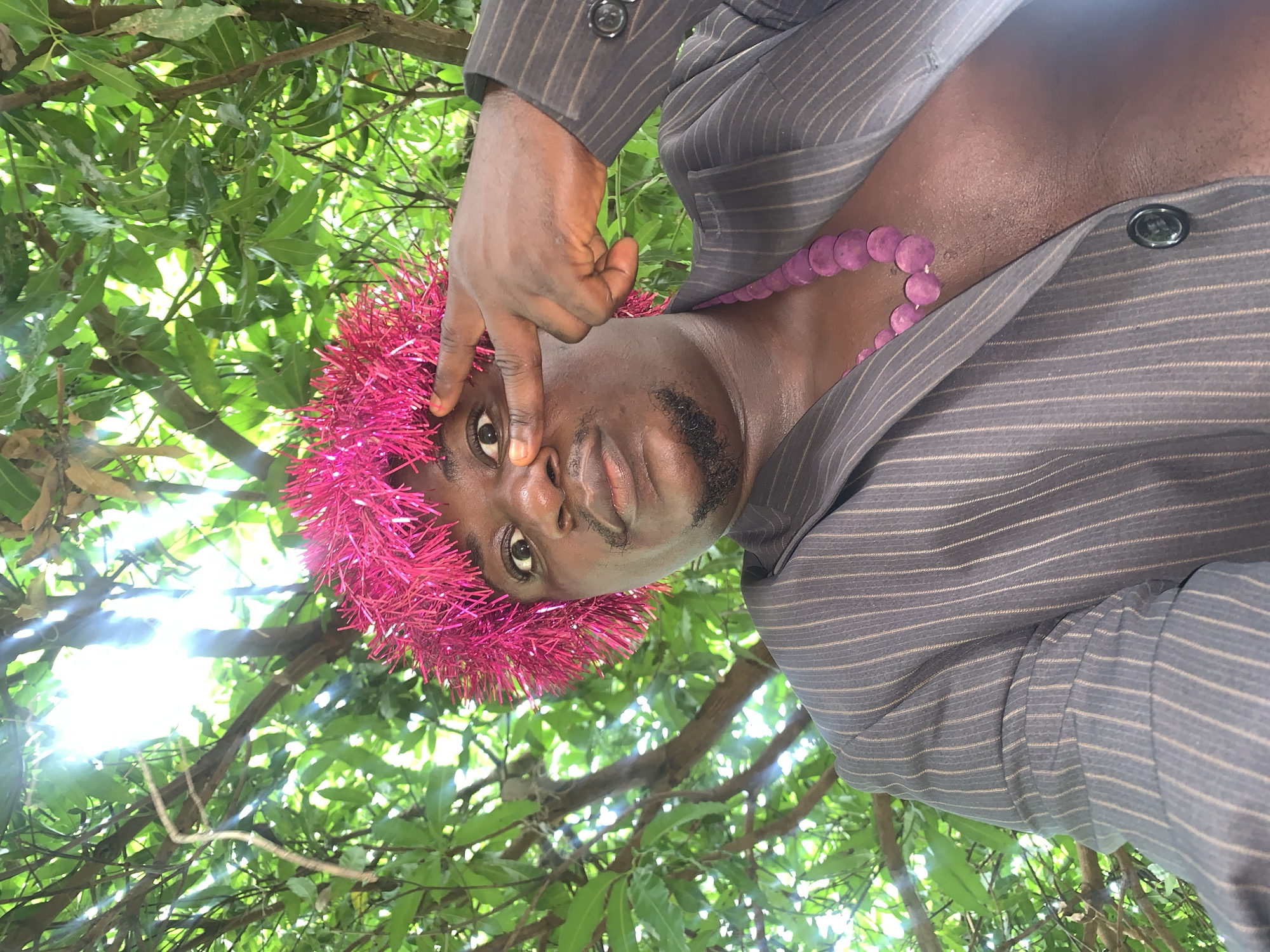 In this close-up, waist-up, outdoor selfie, a person with dark skin and a goatee is positioned centrally, looking directly at the camera. They are wearing a dark pinstripe suit jacket, partially unbuttoned to reveal their bare chest and a thick, purple beaded necklace. A vibrant pink, tinsel-like headdress adorns their head, framing their face. Sunlight streams through the lush green foliage of trees and branches that fill the background, suggesting an outdoor setting in a natural environment within Akwanga, Nigeria. The person's left index finger is extended, touching their left eyebrow in a playful gesture. The overall impression is a candid and bold portrait captured amidst nature.