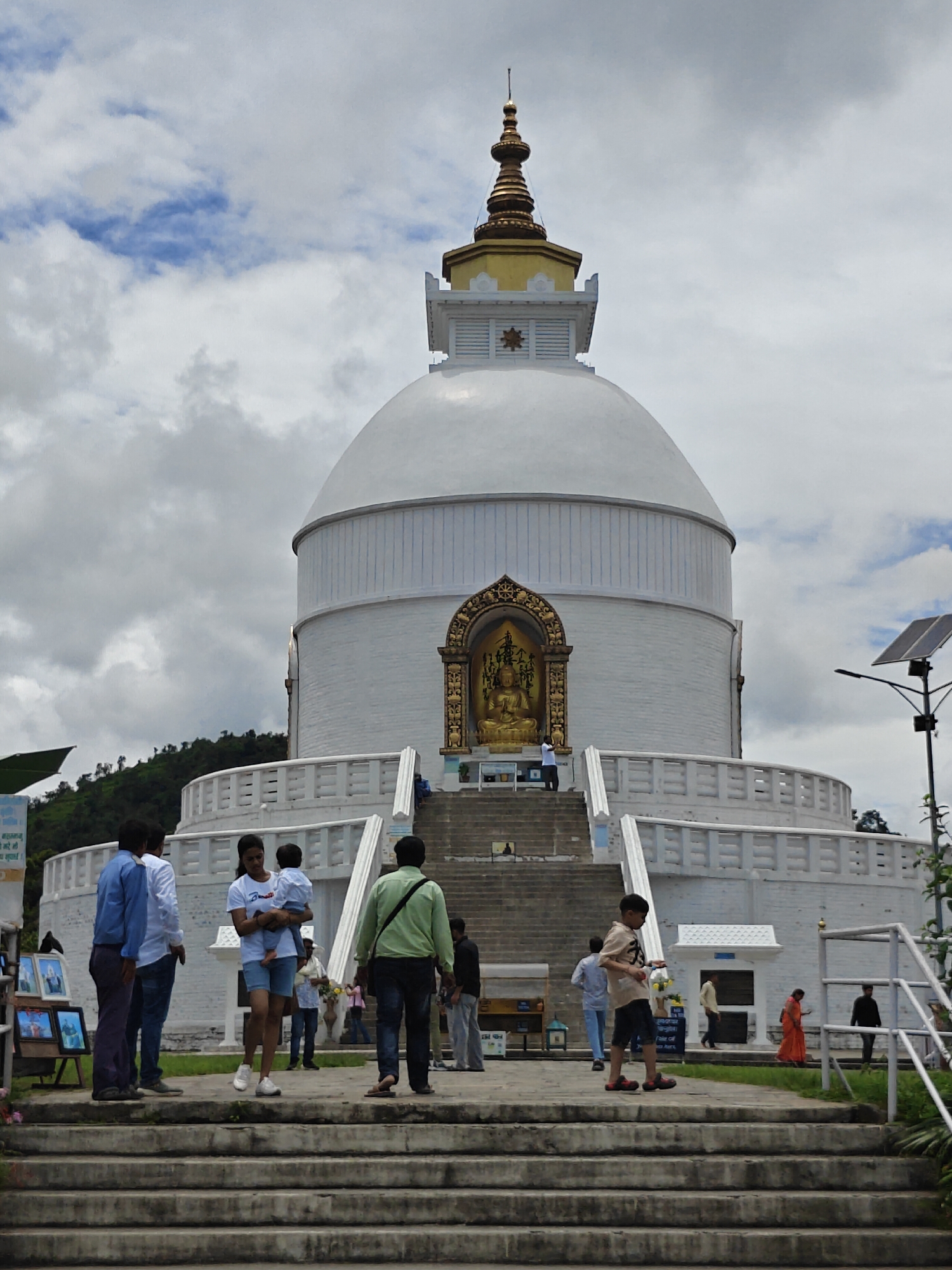 The image shows the World Peace Pagoda in Pokhara, Nepal. The pagoda is a large white structure with a golden spire. It is situated on a hill overlooking the city. In the foreground, a group of people are seen walking up a set of stairs. Some are tourists, while others appear to be locals. The sky is cloudy, and the weather is overcast. The overall scene is peaceful and serene.