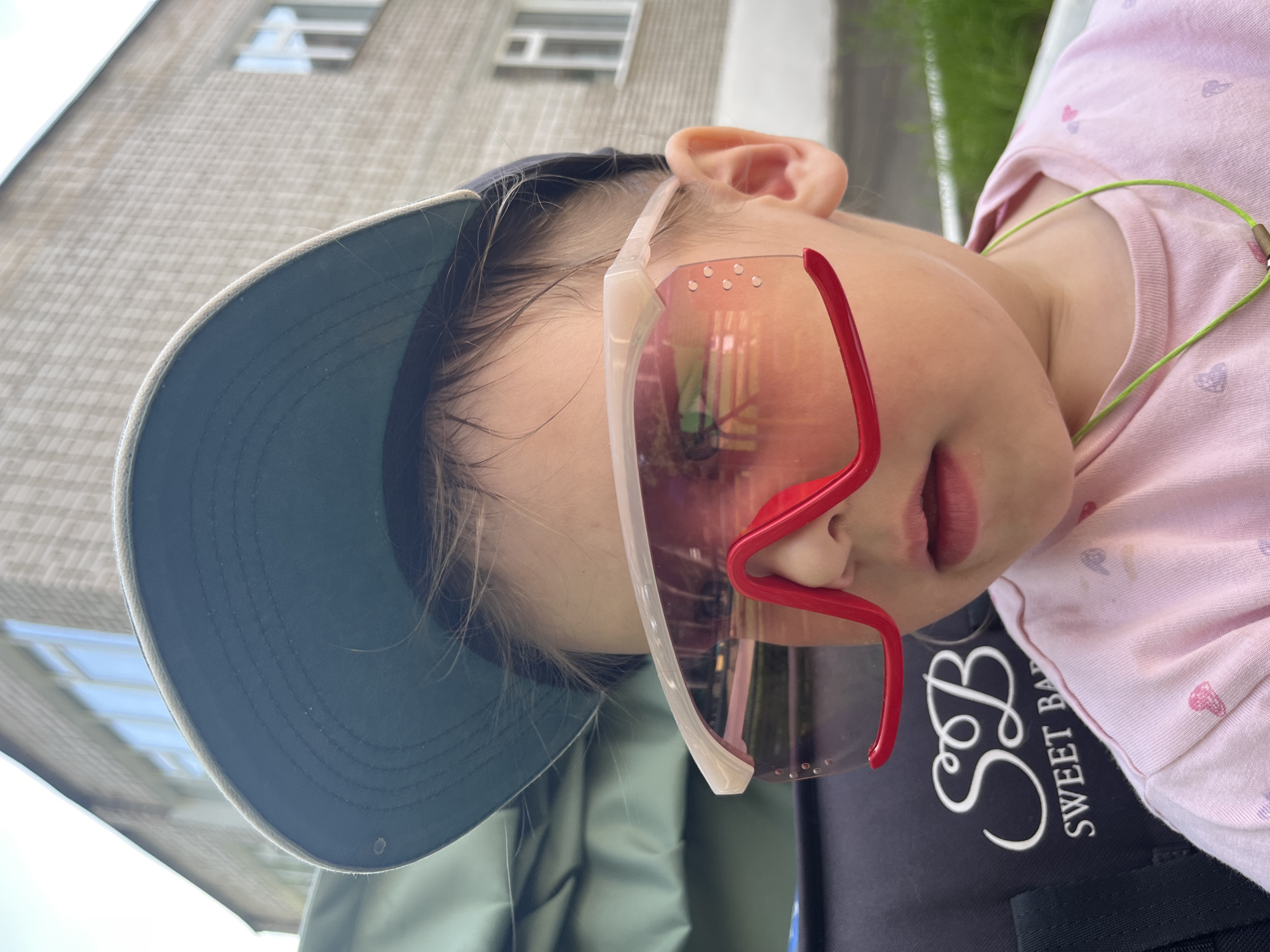 A child wearing a blue baseball cap and oversized white and red sunglasses is resting. The child is wearing a light pink shirt patterned with small hearts. The shirt bears the logo "Sweet Baby." The background includes a gray building with windows, suggesting an urban environment. The scene is likely outdoors during daylight hours. Based on the location provided (Chaikovsky, Russia), the urban environment is assumed to be within this city.
