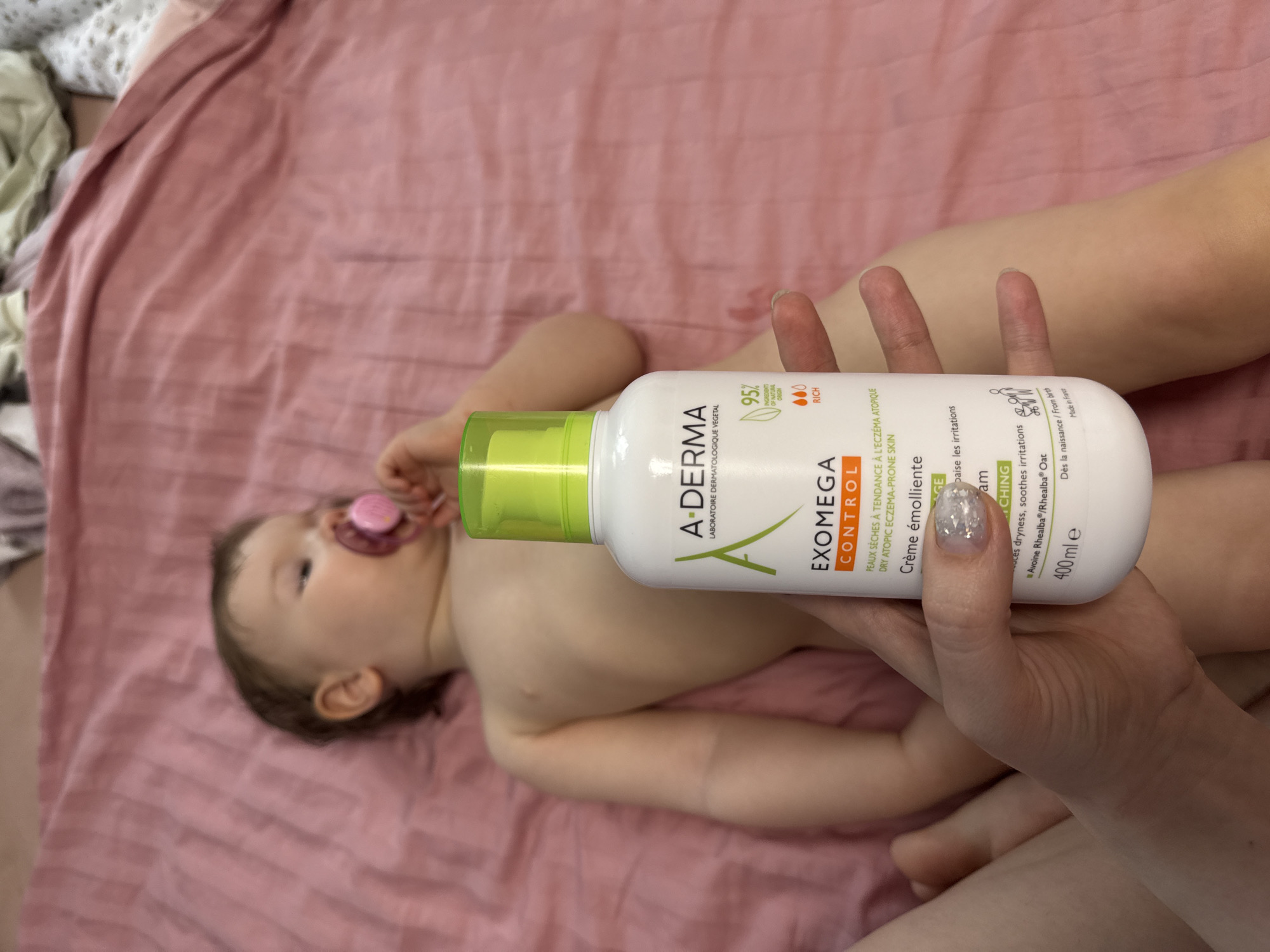 An overhead indoor shot shows a baby lying on its back on a textured pink sheet or blanket. The baby, with dark hair and fair skin, has a pink pacifier in its mouth and appears calm, looking towards the left side of the frame. An adult's hands are visible on the right, holding a white and bright green bottle of A-Derma skincare product.
The label on the bottle is clearly legible, identifying the product as "A-DERMA LABORATOIRE DERMATOLOGIQUE VEGETAL EXOMEGA CONTROL Crème émolliente." It states the cream is for "PEAUX SÈCHES À TENDANCE À L'ECZÉMA ATOPIQUE" (Dry Atopic Eczema-Prone Skin), "baise les irritations" / "soothes irritations," and contains "Avoine Rhealba®/Rhealba® Oat." Additional details include "95% INGREDIENTS OF NATURAL ORIGIN," "RICH," "Dès la naissance / From birth," "Made in France," and a volume of "400 ml e." One of the adult's fingernails is painted with silver glitter polish. The scene, likely taking place in a home in Moscow, Russia, depicts a routine care moment, possibly for moisturizing the baby's skin.