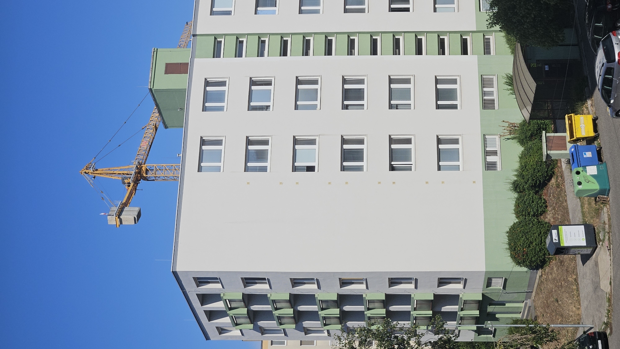 The image shows a tall, light gray residential building with green balconies and accents, set against a clear blue sky. A large yellow construction crane is visible extending over the top of the building. In front of the building, there are several colorful recycling bins, including green, blue, and yellow ones, as well as a dark gray bin with labels. A small, covered structure resembling a bus stop or shed is situated to the right of the recycling bins. There are also trees and bushes around the building and in the foreground, with a street and parked cars visible at the bottom of the image. The scene appears to be in a residential area in Bratislava, Slovakia, possibly during daytime given the bright sunlight and shadows.