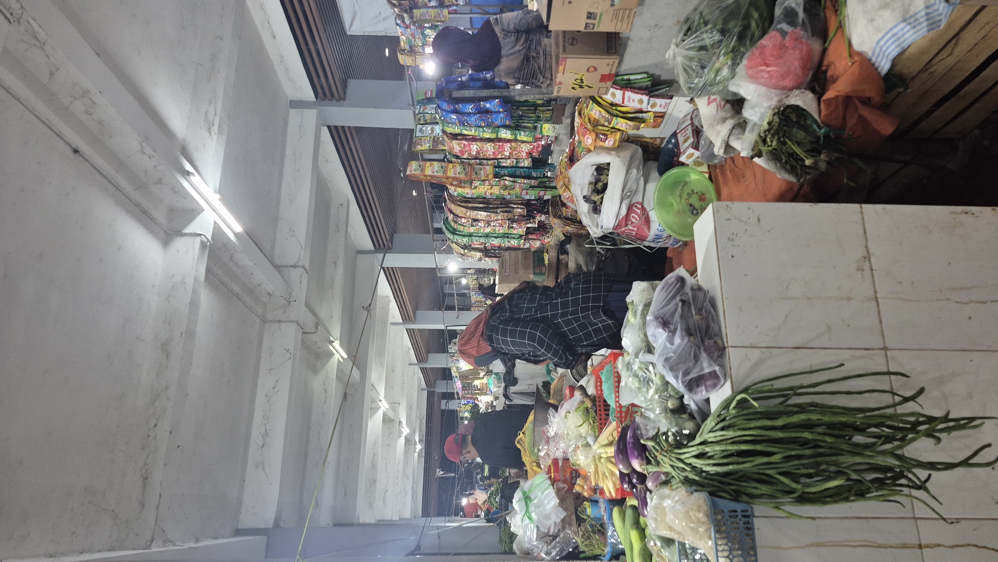 The image depicts a bustling indoor market scene, likely a traditional wet market, in Bojonegoro, Indonesia. In the foreground, a variety of fresh produce is displayed, including long green beans, eggplants, and cucumbers, some of which are bagged or contained in baskets. The stall is constructed with white tiled surfaces. Behind the produce, several individuals are visible. One person, wearing a black and white checkered jacket and a red headscarf, appears to be a vendor or shopper, looking down at the goods. Another person, wearing a red cap and black attire, is seen from the back, also seemingly engaged in market activities. Further back, more people are visible amidst stalls laden with packaged goods, predominantly spice sachets or instant food products hanging in rows. The market appears to be covered by a concrete ceiling with exposed beams and fluorescent lighting fixtures, suggesting a permanent market structure. The overall atmosphere is one of active commerce and community interaction within a lively Indonesian marketplace.