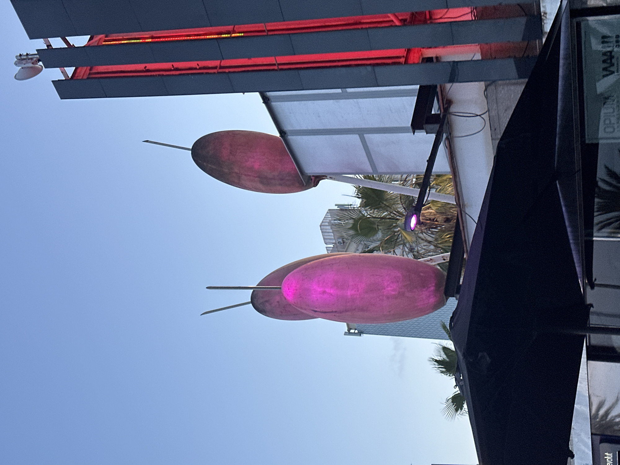 The image depicts a scene in Barcelona, Spain, featuring large, pink, oval-shaped sculptures that appear to be illuminated from within or by external lighting. These sculptures are positioned outdoors, against a backdrop of a clear blue sky at dusk. To the right of the sculptures is a modern building facade with vertical red lighting. In the foreground, a dark umbrella is visible, partially obscuring the lower portion of the frame. In the background, glimpses of other buildings and palm trees suggest an urban environment. There are no people visible in the image.