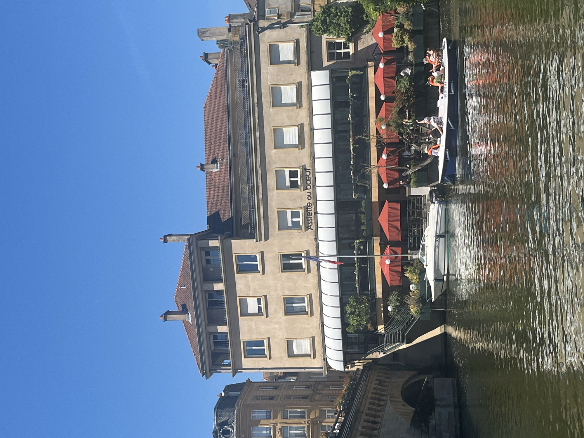 The image depicts a waterscape alongside a building in Metz, France. The building, with a beige facade and red-tiled roof, features a sign that reads "Assiette au Boeuf." A French flag is displayed. A retractable awning covers an outdoor seating area with red umbrellas. Two boats are in the water. One boat contains several people wearing orange life jackets. The water reflects the building and sky. A stone bridge is visible in the foreground. The sky is clear and blue.