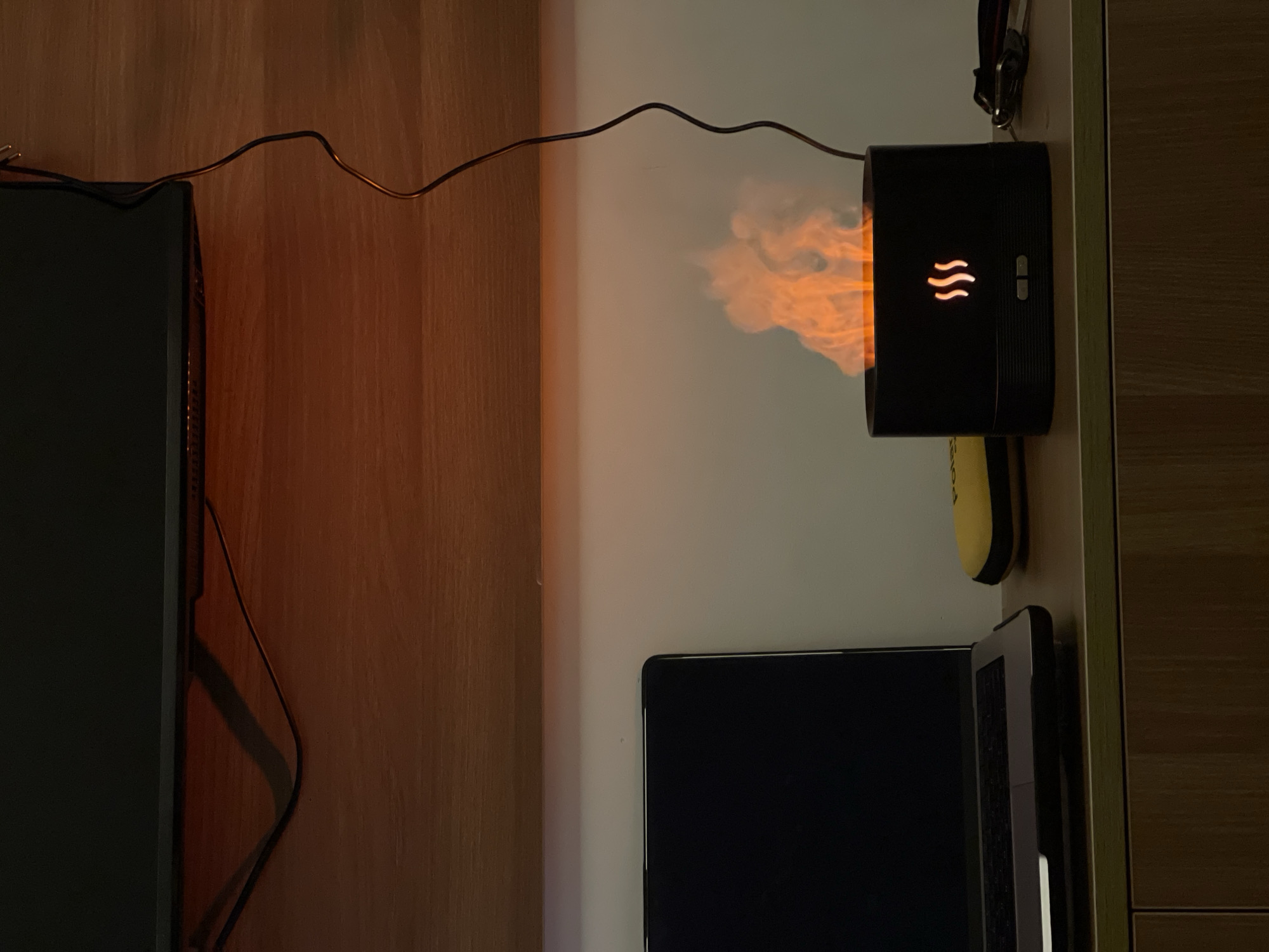 This is a close-up, eye-level, indoor shot of a desk setup, likely in an office or home office in Đà Nẵng, Vietnam, given the context of similar images from the area. The primary subject is a black, cylindrical flame humidifier, which is emitting a realistic artificial flame effect with orange and yellow hues. The humidifier has two small buttons on the front and a stylized flame icon illuminated above them. To the left of the humidifier is a silver laptop, open with its screen dark. Behind the humidifier and to its right, are some small items including a yellow and black case and part of a keychain with metal clips. The objects are placed on a light-colored wooden desk surface. Above the desk is a wooden shelf, and above that, a dark, flat surface, possibly a monitor or the underside of a cabinet. A black power cord for the humidifier snakes from the device upwards and out of frame. The lighting is dim, with the artificial flame and the illuminated icon on the humidifier providing most of the light, casting warm tones on the surrounding surfaces and creating shadows.