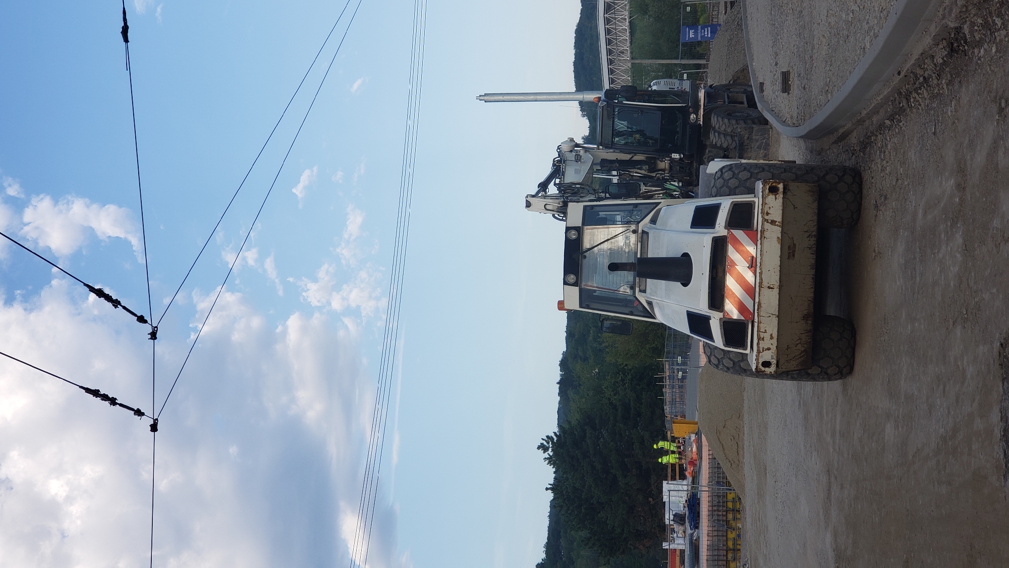 In Prešov, Slovakia, a construction site is captured in the image under a partly cloudy sky with overhead power lines. The foreground is dominated by a white construction vehicle with large, rough tires and distinctive orange and white striped markings on its rear. To the right and slightly behind it, a black excavator is partially visible. Further in the background, two construction workers in yellow safety vests are seen near what appears to be the foundation of a structure. The environment includes piles of gravel and sand, as well as fencing and other construction materials. The presence of overhead power lines suggests this might be a location where a tram or trolleybus system operates or is under construction.