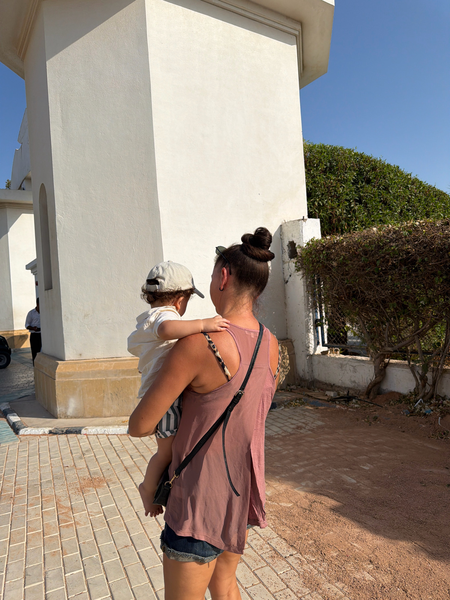 A woman, viewed from behind, carries a toddler in her left arm while walking on a paved walkway. The woman has dark hair styled in a bun and is wearing a pink tank top and denim shorts. The toddler is wearing a light-colored shirt and a cap. Behind them, a light-colored building with architectural details and a hedge are visible. The ground is a mixture of paved tiles and reddish-brown dirt. The scene appears to be in a sunny outdoor location, possibly Sharm El Sheikh, Egypt, given the architectural style and vegetation. A person in a white shirt is visible in the background, standing near the building.
