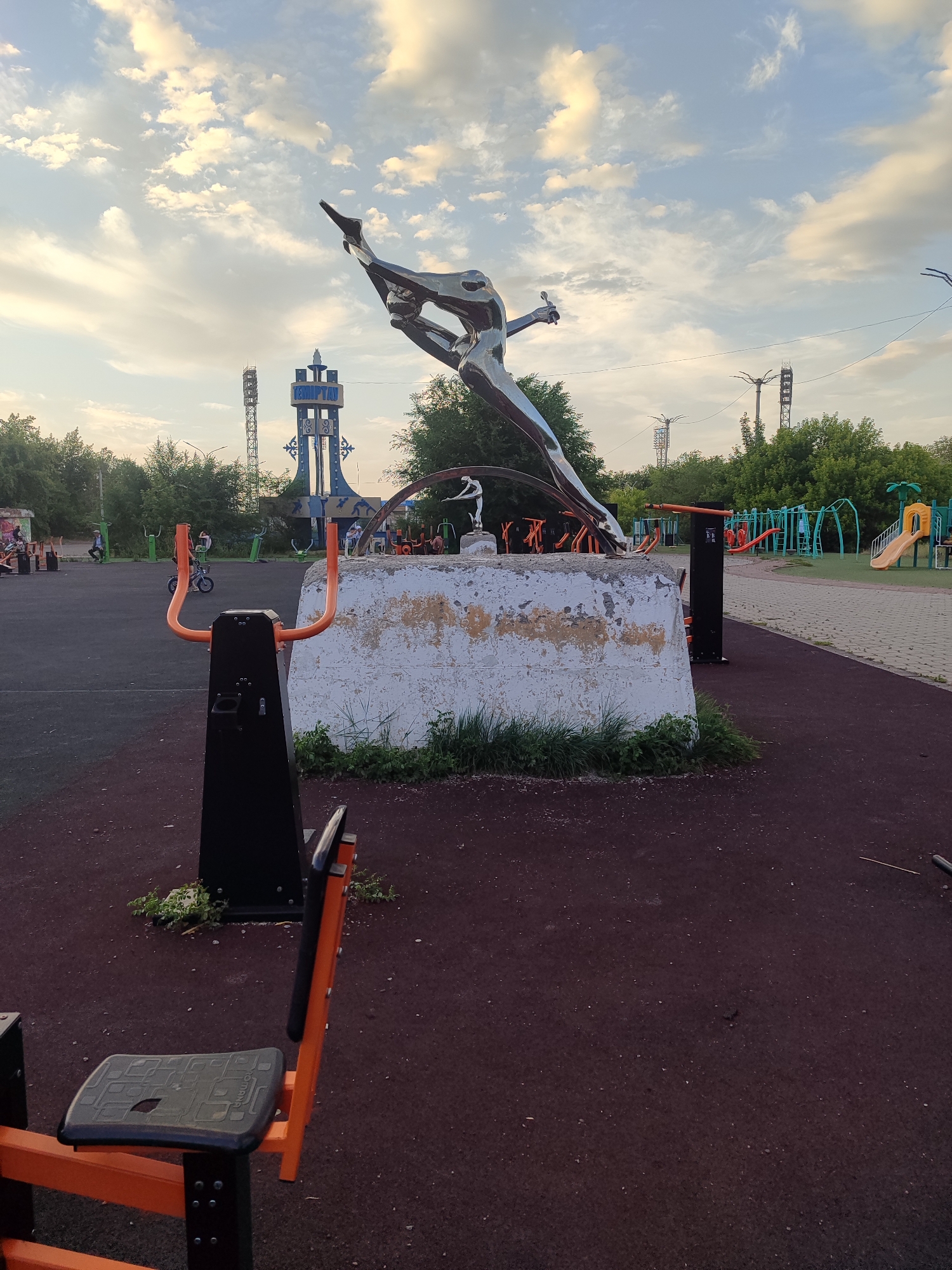 The image depicts an outdoor sports and recreation area in Temirtau, Kazakhstan. Dominating the foreground is a modern outdoor exercise machine with orange handlebars and a black seat. Behind it, a large, weathered concrete pedestal supports a gleaming metal sculpture of a person in a dynamic, athletic pose, possibly representing a swimmer or diver. The sculpture is highly reflective, catching the light from the sky.
In the mid-ground, the sports area is furnished with additional exercise equipment, some in green and orange, and a children's playground with slides and climbing structures is visible further in the background. A prominent blue and white tower with ornate detailing stands tall in the distance, bearing the Cyrillic text "Темпт". Several people are scattered throughout the area, some engaged in activities like riding a small motorcycle, while others appear to be observing or resting.
The time of day appears to be late afternoon or early evening, with soft, diffused sunlight illuminating the scene and casting gentle shadows. The sky is a mixture of blue and white clouds, suggesting fair weather. The overall atmosphere is one of active community use and leisure, with a blend of modern recreational facilities and artistic public installations. The location is clearly an urban park or public square designed for physical activity and social gathering.