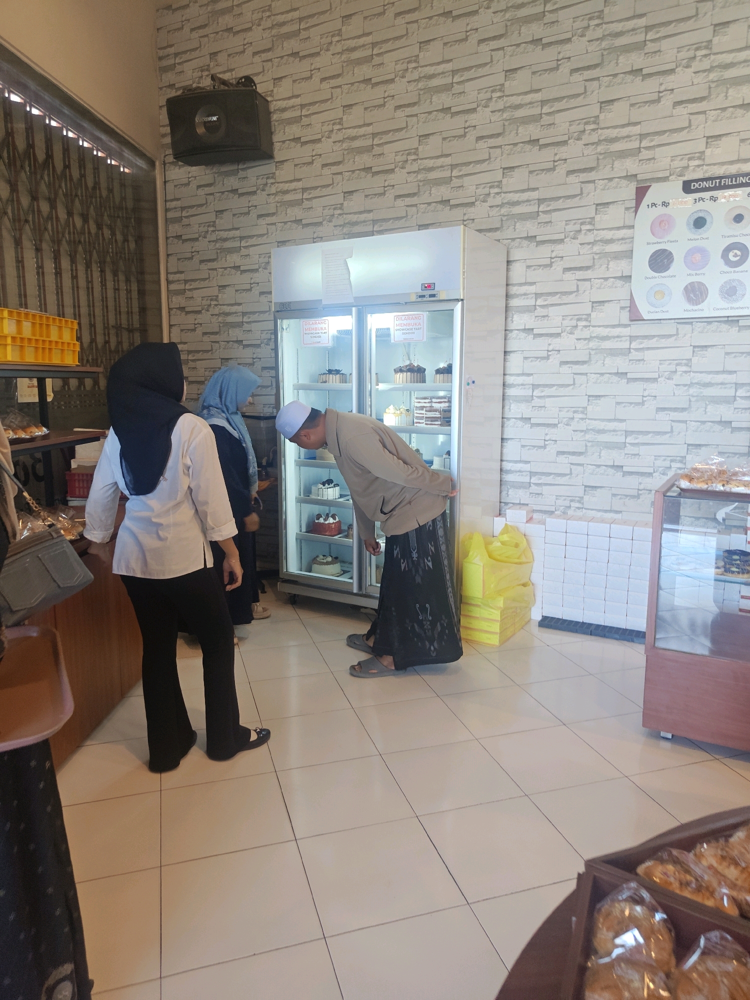 This image depicts the interior of a bakery in Situbondo, Indonesia, on a sunny day. A man wearing a light brown shirt, black pants, and a white skullcap is leaning over to examine cakes in a refrigerated display case. Two women, one in a black hijab and white shirt, the other in a blue hijab, stand nearby, also looking at the display. The display case, labeled "GEA," contains several cakes, including a tiered chocolate cake and other pastries. To the left of the display case, there are shelves with yellow food crates and bags of bread. In the background, a brick-patterned wallpaper adorns the wall, and a black speaker is mounted near the ceiling. Boxes and bright yellow shopping bags are stacked on the floor to the right of the display case. A glass display case filled with baked goods is visible on the far right. The floor is tiled with light-colored squares.