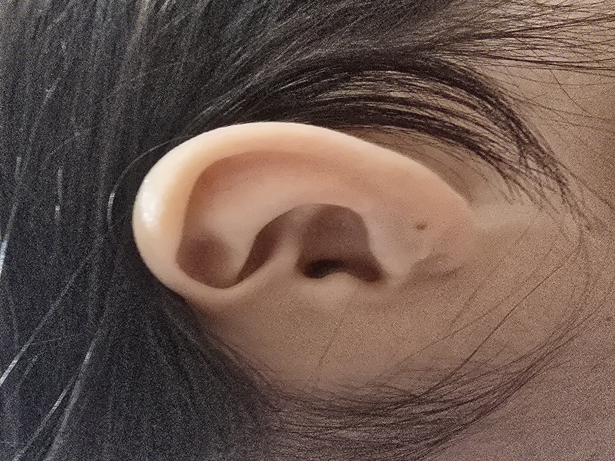 The image is a close-up of a human ear, framed by dark hair. The ear is light-skinned with visible contours and a small mole. The hair is dark and appears to be somewhat messy, with individual strands catching the light. The background is out of focus and consists of dark tones. There is no indication of a specific location in the city of Ulan-Ude, Russia, or any discernible environmental context. The scene is static and lacks any action or interaction.