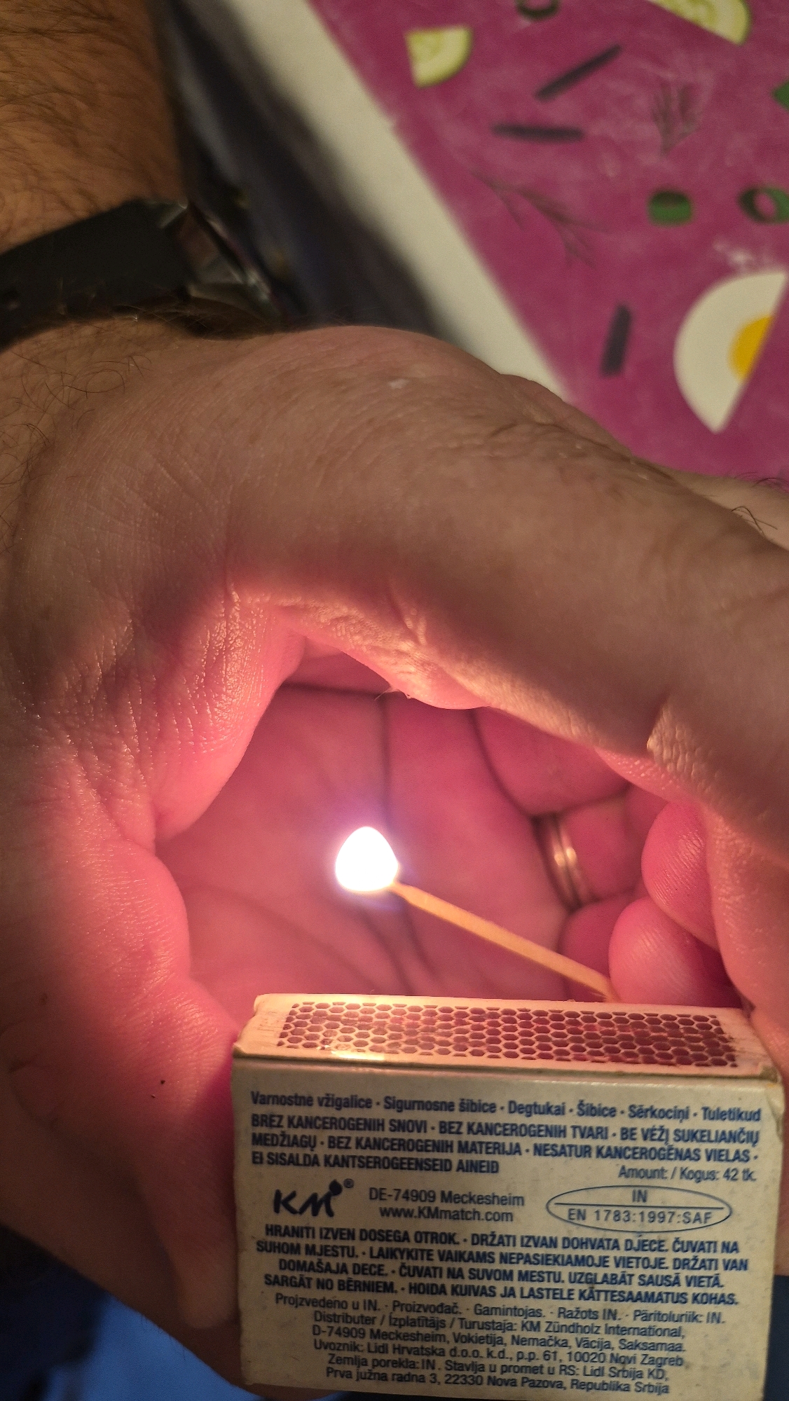A close-up image shows a person holding a lit match over an open box of matches. The match head is glowing brightly, and the flame is visible. The box of matches has text printed on it, suggesting it is a safety-related product with warnings and information in multiple languages. The background is out of focus but appears to be a brightly colored surface with some food items depicted, possibly a placemat. The scene is indoors, with artificial lighting creating a warm glow. There are no discernible geographical markers or clues to indicate the location is in Krauja, Latvia.