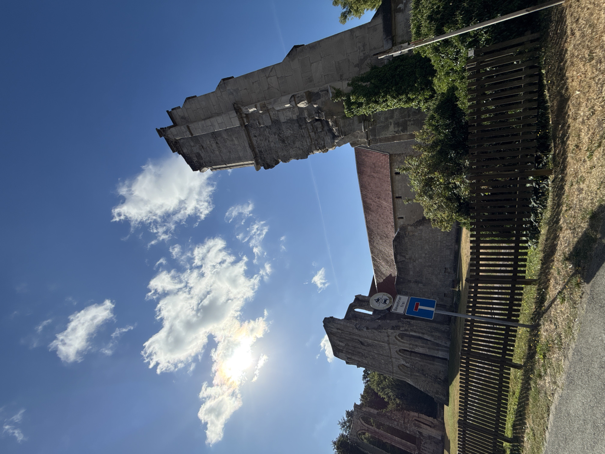 The image shows a view of the ruins of the Walkenried Abbey in Walkenried, Germany, on a clear, sunny day. The sky is a vibrant blue with scattered white clouds, and the sun is shining brightly, casting long shadows and creating lens flare. In the foreground, a wooden fence runs horizontally across the frame, separating a dry, grassy area from the abbey ruins. To the left of the fence, a paved road is visible.
The abbey ruins are constructed from stone and appear to be partially preserved and partially in disrepair. A prominent tall, stone tower, leaning slightly, is a central feature. To the left of this tower, sections of arched windows and stone walls are visible, with greenery growing on and around the structures. A building with a red tiled roof is situated behind the ruins, suggesting a more intact section of the historical site. A traffic sign with a motorcycle, a car, and text reading "Bewohner frei" (residents free) and a blue sign with a white horizontal bar indicating a dead end is positioned near the fence, leaning slightly. Lush green trees and bushes surround the abbey ruins. The overall scene depicts a historical landmark bathed in bright sunlight.