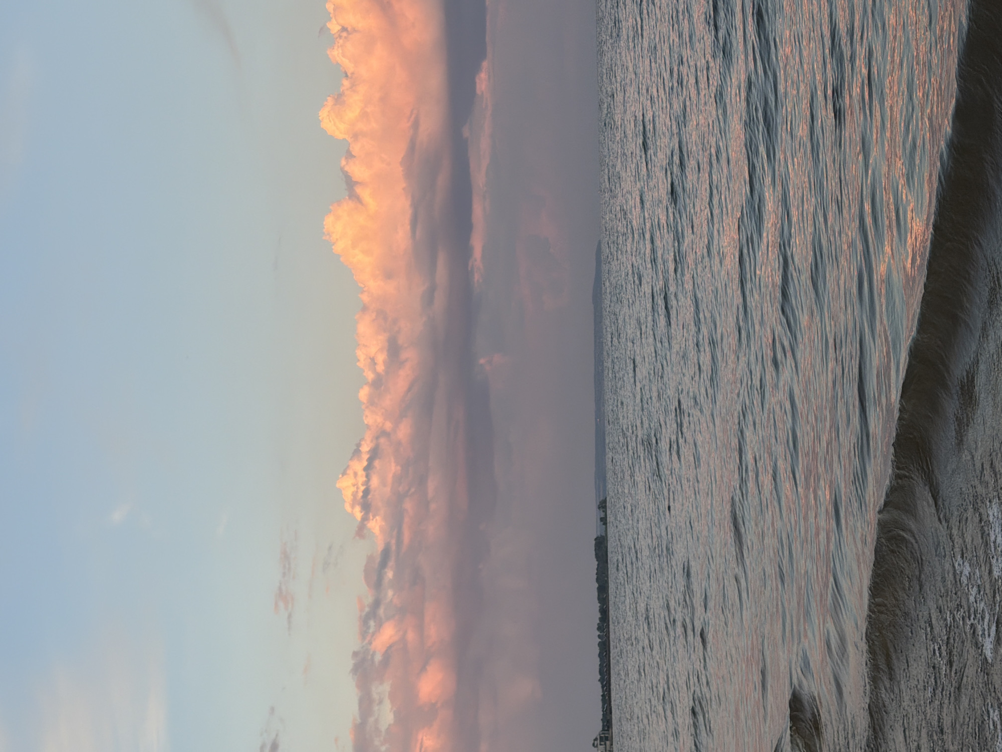 The image depicts a serene seascape at dusk, possibly from the shore of Laishevo, Russia. The sky is a soft gradient of pale blue at the top transitioning to a hazy lavender near the horizon. A band of fluffy clouds, illuminated with warm pink and orange hues by the setting sun, stretches across the middle of the sky. Below the clouds, a distant, dark, and low-lying landmass is visible, with what appears to be a developed shoreline featuring structures and trees. The water in the foreground is calm, with gentle ripples creating a textured surface that reflects the muted colors of the sky. The overall impression is one of tranquility and the soft beauty of a coastal sunset. There are no people or discernible actions taking place.
