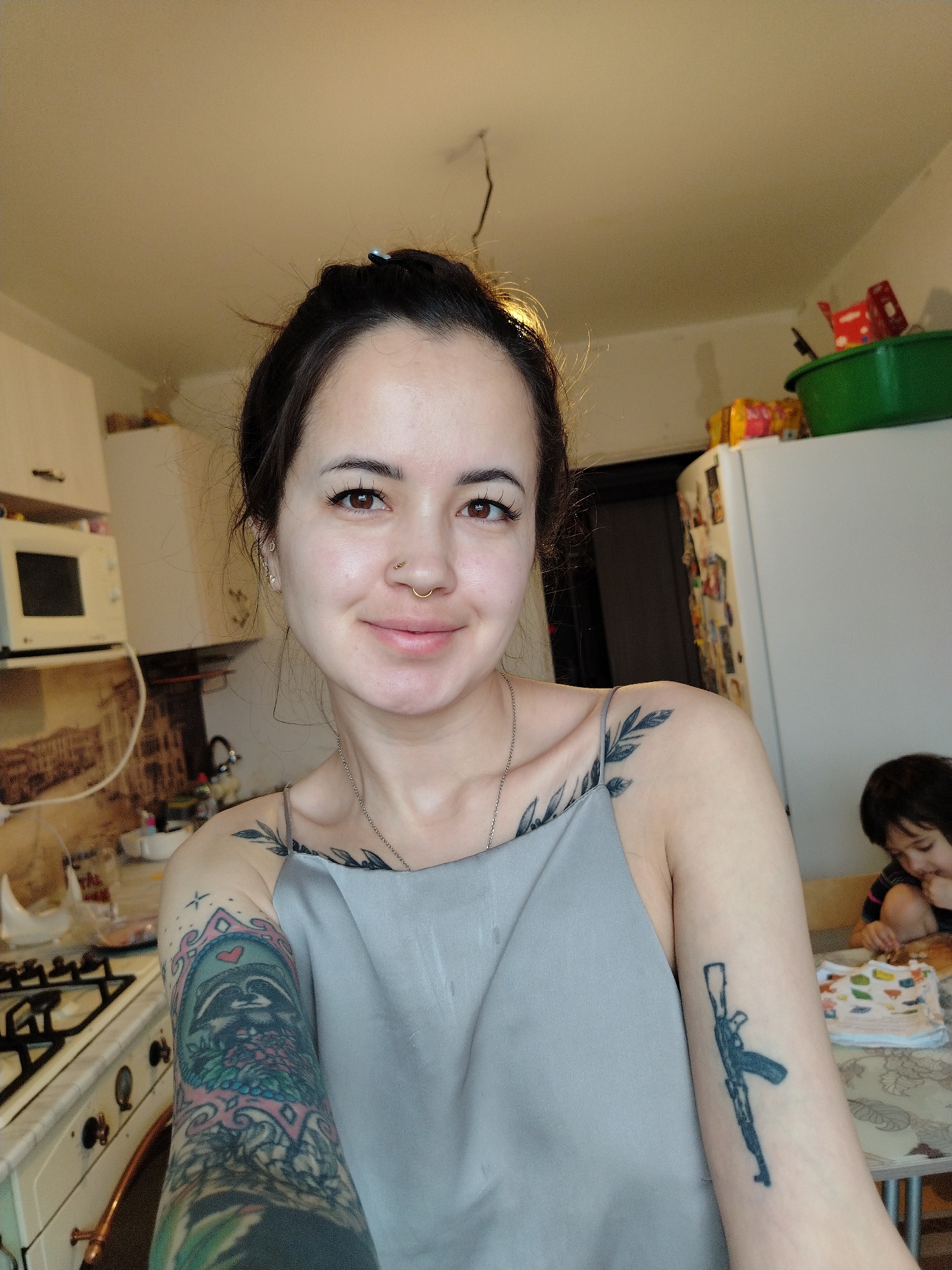 A young woman takes a selfie in what appears to be a kitchen. The kitchen has white cabinets, a microwave, and a gas stove. A small child sits at a table in the background. The woman has several tattoos on her arms, including one of a raccoon and another of a firearm. The specific location is not determinable from this image.