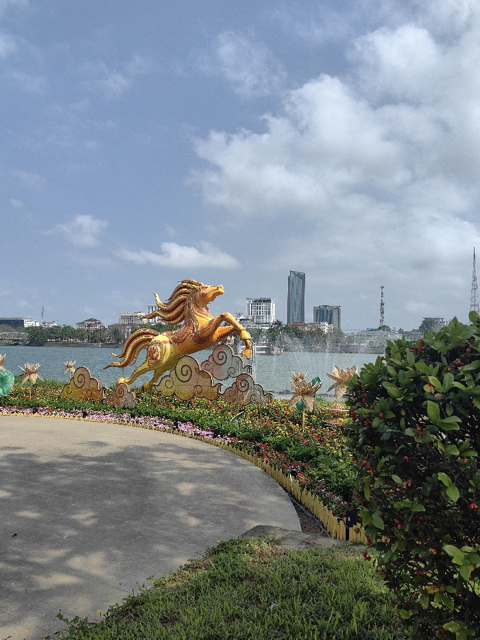 The image captures a daytime scene within a park area adjacent to a body of water in Thuận Hóa Ward, Vietnam, under a partly cloudy sky. A prominent, large, golden-orange equestrian statue, depicted as a horse mid-leap on stylized white clouds, occupies the mid-ground. This statue is surrounded by a meticulously landscaped flowerbed featuring various colorful plants and decorative golden lotus-shaped ornaments, bordered by a low, yellow picket fence-like barrier. A curved, light-grey concrete pathway is visible in the lower-left foreground, displaying dappled shade patterns. To the right of the pathway, patches of green grass and a dense green shrub with small red berries are present. Beyond the landscaped area, a wide body of water extends, exhibiting a visible water fountain or spray in the mid-distance. The background features a city skyline across the water, including a tall, slender, dark-colored skyscraper, several lighter-colored multi-story buildings, and a distant metal lattice communication tower on the far right. A bridge structure is faintly visible on the far left horizon. No individuals are depicted within the scene.