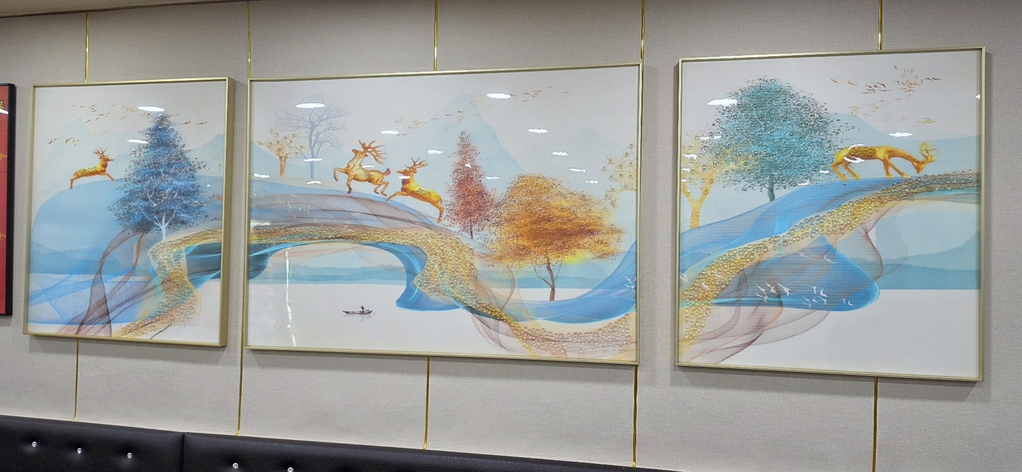 The image displays three modern art pieces hung on a textured wall with vertical gold fixtures. The artwork features a stylized landscape with a serene, light-colored background. Each panel depicts a flowing, abstract representation of a river or path in shades of blue and gold. Interspersed are decorative trees with textured foliage in blues, oranges, and yellows. Golden deer figures are positioned on the abstract paths in various poses, suggesting movement and grace. The scene is evocative of a natural environment with elements of fantasy. The wall behind the artwork is a neutral, textured beige, and the gold fixtures run from the top of the frames down to an unseen area below. The lighting appears to be overhead and casts a slight sheen on the artwork. There are no people visible in the image. This artwork is likely displayed in a commercial establishment or a home interior in Cheongju-si, South Korea.