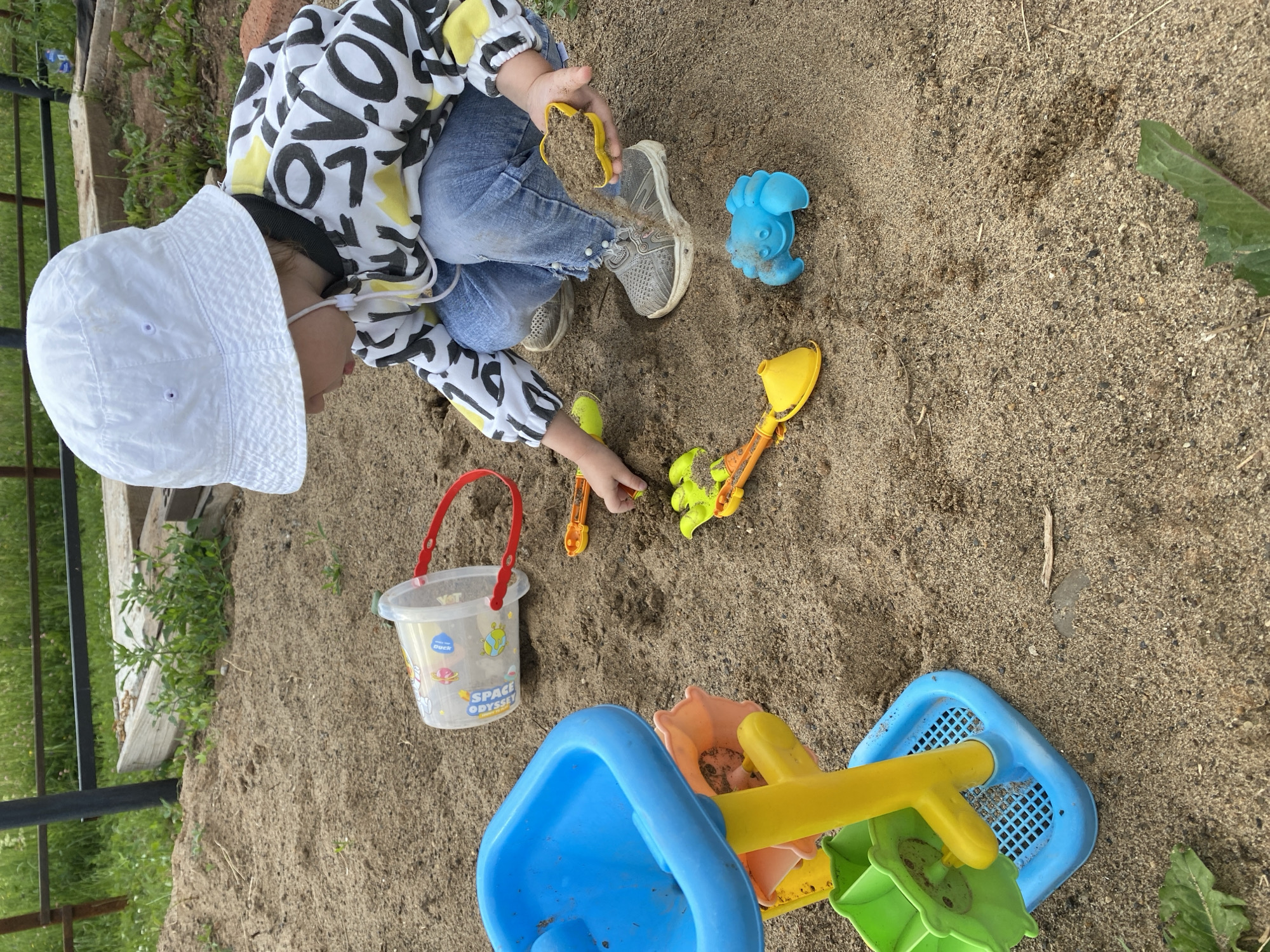 A young child, wearing a white bucket hat and a long-sleeved shirt with a black, white, and yellow graphic pattern, along with blue jeans, is playing in a sandbox. The child is crouched down and appears to be scooping sand with a yellow flower-shaped mold. Several other sand toys are scattered around, including a blue water wheel toy, a yellow shovel, a green rake, and a blue crab-shaped mold. A transparent bucket with a red handle is also present, lying on its side in the sand. The environment is outdoors, with sandy ground and green vegetation visible in the background, suggesting a park or a backyard. The lighting is natural, indicating daylight.