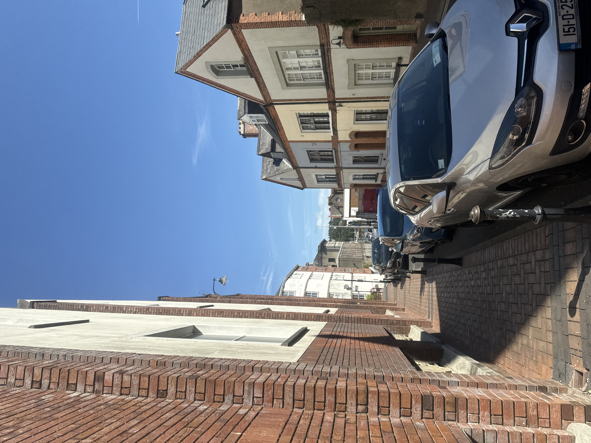 The image depicts a street scene in Waterford, Ireland, bathed in sunlight under a clear blue sky. The perspective is from a low angle looking down a brick-paved sidewalk lined with parked cars and residential buildings. The buildings on the left are predominantly red brick, with a lighter colored section on the right side of the image featuring multiple stories with dormer windows and red brick accents around the windows and rooflines. The buildings on the right are varied in color, including shades of cream, yellow, and a pale blue-grey, with large windows. Several cars are parked along the street, with the most prominent being a silver Renault hatchback in the foreground. The license plate of the silver car is visible and reads "151-D-25". A lamppost is visible on the left side of the sidewalk, casting a shadow. In the distance, more buildings and trees are visible, suggesting an urban or semi-urban environment. The overall impression is a quiet, sunlit street in a residential area.